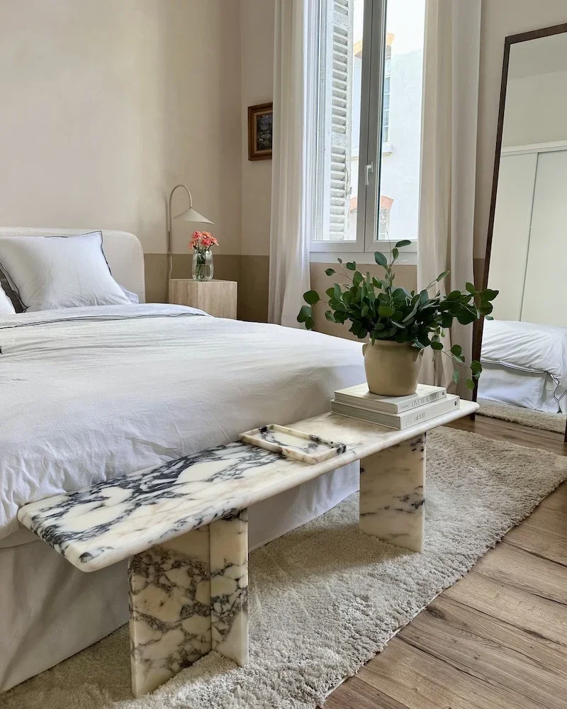A cozy bedroom with a white bed, a marble bench at the foot of the bed, a potted plant, a nightstand with pink flowers, a mirror, and a window with white curtains.