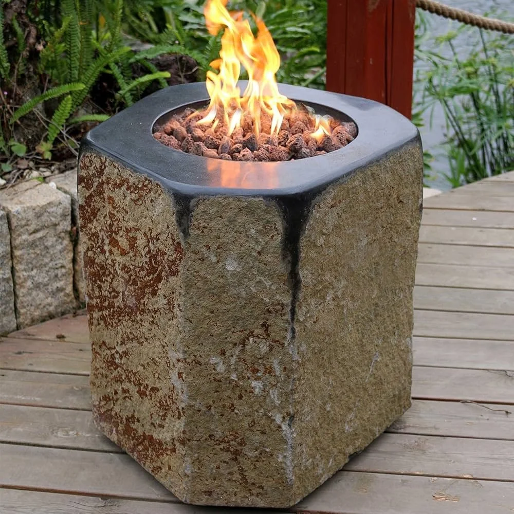 A cube-shaped outdoor fire pit made of stone with a metal top, burning with flames on a wooden deck near a lake and greenery.