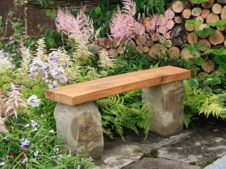 Wooden garden bench with stone supports in a lush garden with various green plants and pink flowers, and a woodpile in the background.