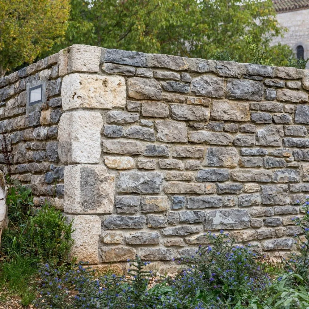 Stone wall with mixed large beige and gray stones, surrounded by greenery and purple flowering plants.
