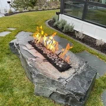 Outdoor fire feature built into a large stone bench with a controlled flame, situated on a lawn near a house.