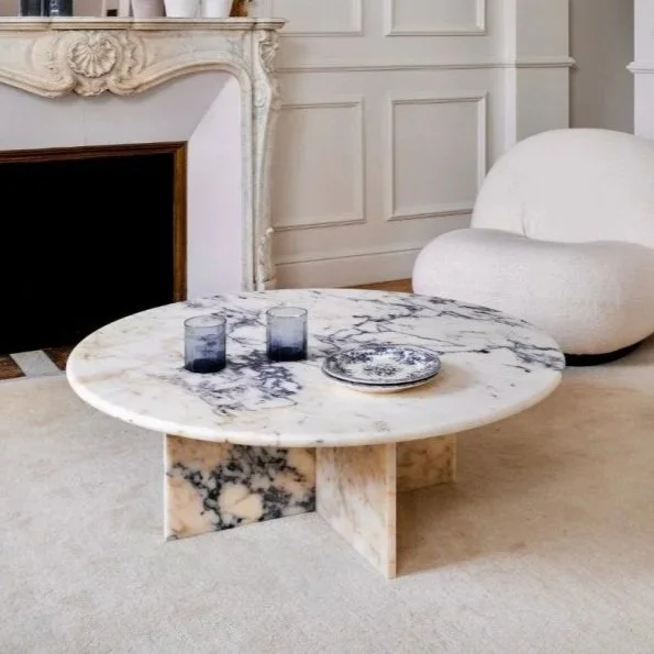 A marble coffee table with two dark-colored glasses and a decorative plate, situated in a living room with a cream-colored armchair, decorative wall paneling, and a white fireplace with ornate detailing.