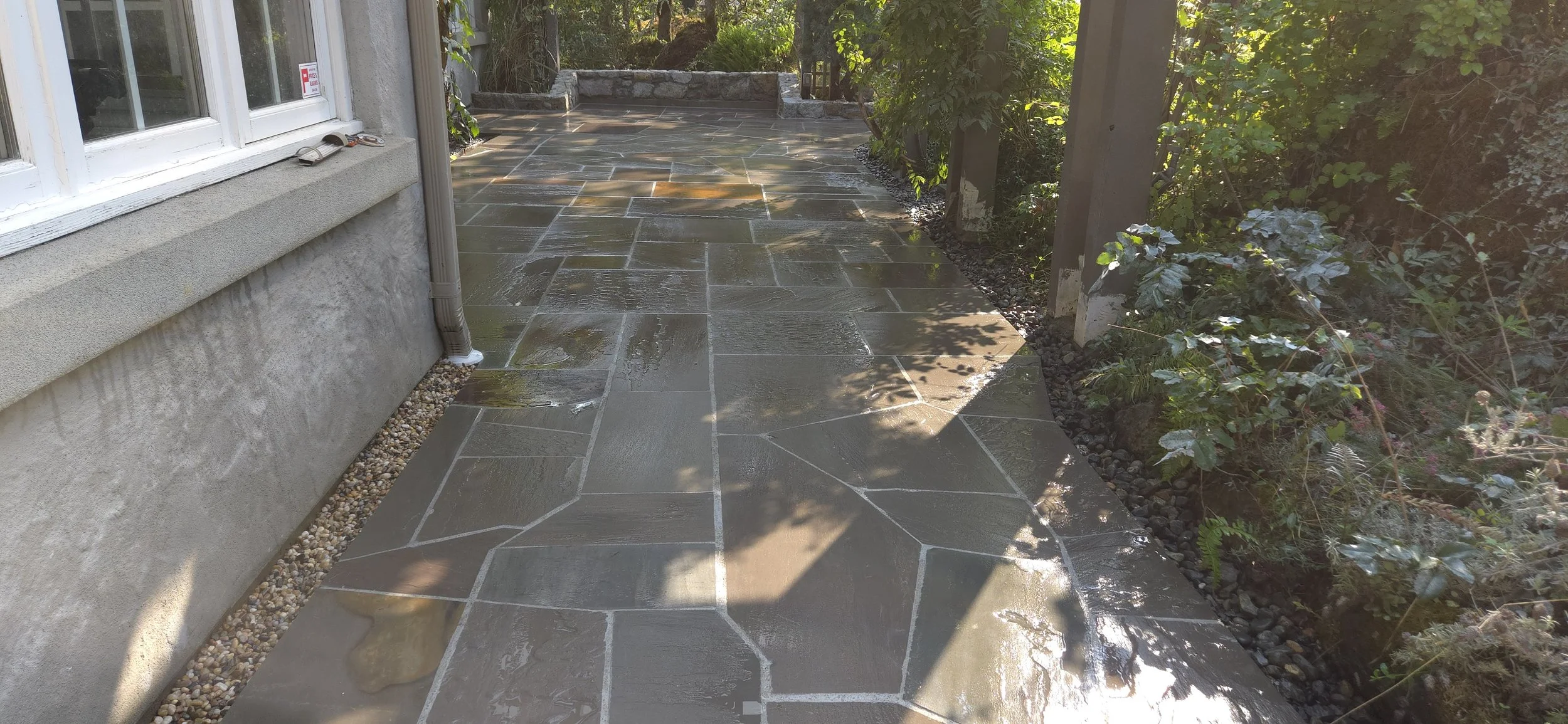 A wet stone patio next to a house with a window on the left, a rain gutter downspout, and a garden with plants and a wooden fence on the right, with sunlight and shadows.