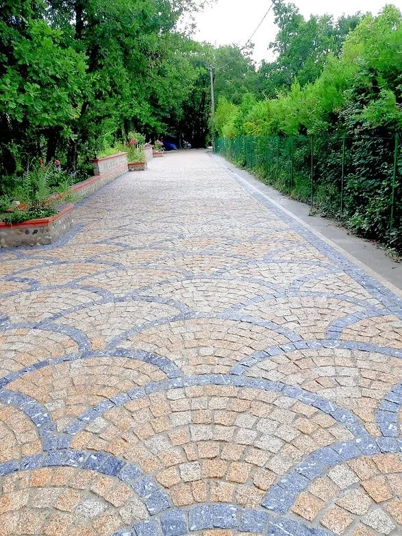 Cobblestone pathway lined with green trees and bushes on both sides, with a small brick wall on the left side and a fence on the right side.