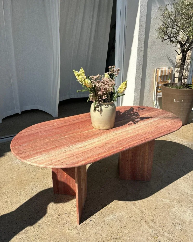 A travertine table with a vase and flowers on it, set outside with with white curtains and a potted tree in the background