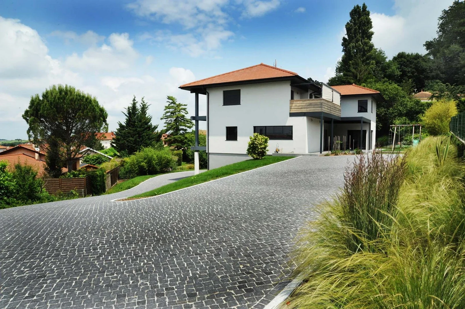 Modern white house with a red tile roof, located on a sloped driveway made of interlocking black stones, surrounded by greenery, trees, and shrubs.