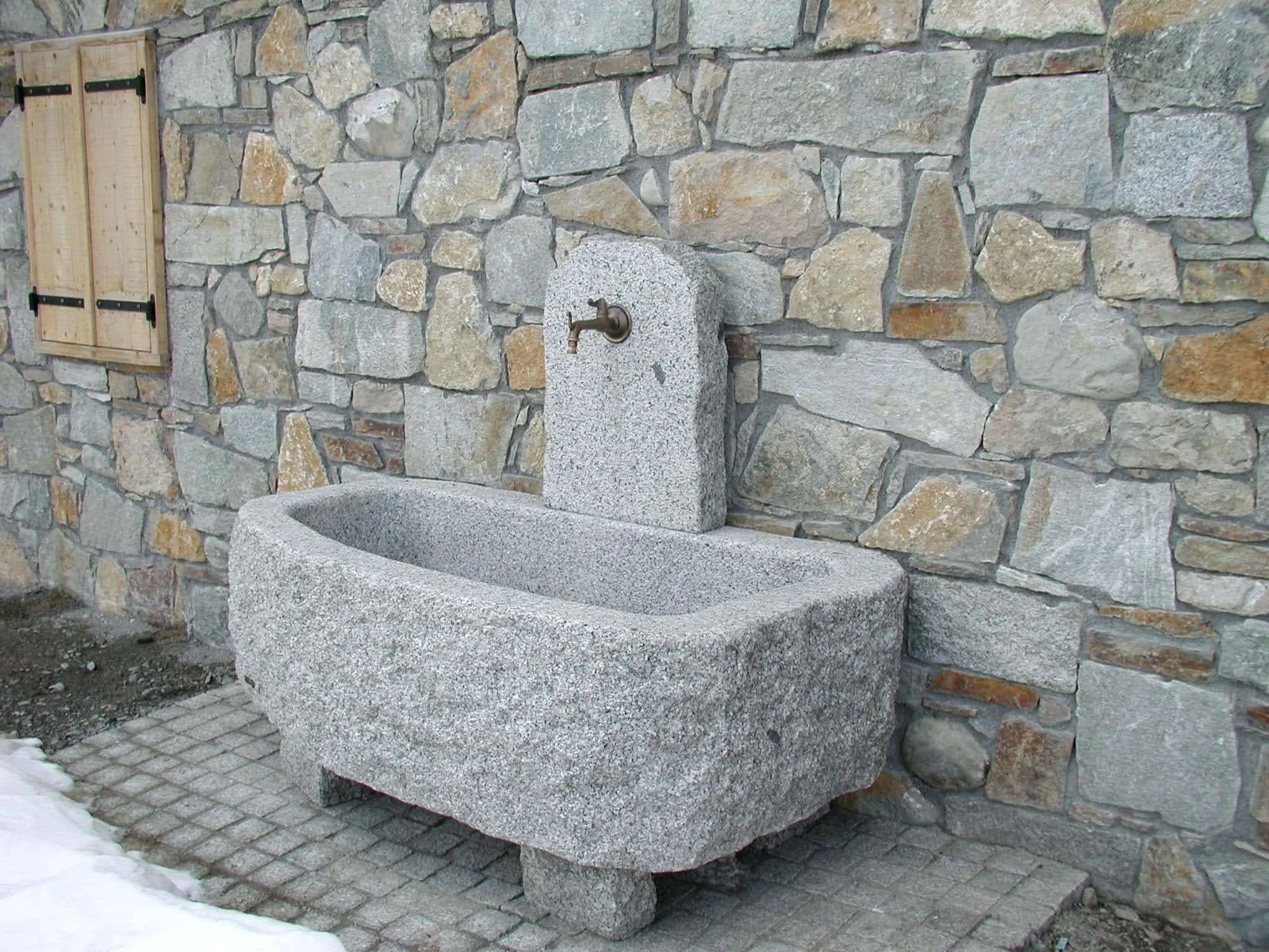 An outdoor stone water basin with a wall-mounted faucet against a stone wall, with some snow on the ground.