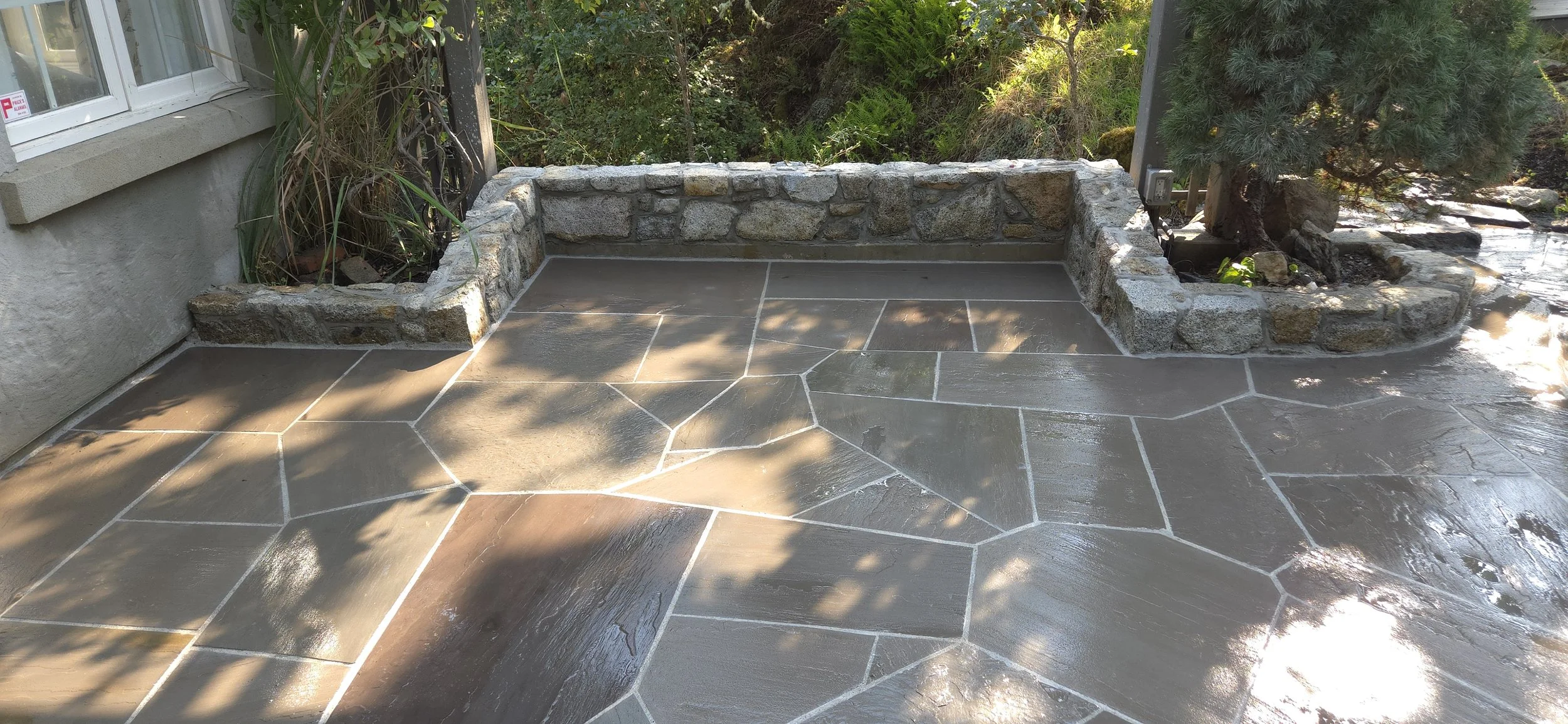 A stone-edged garden bed on a stone patio with a small pine tree and plants, partially shaded.