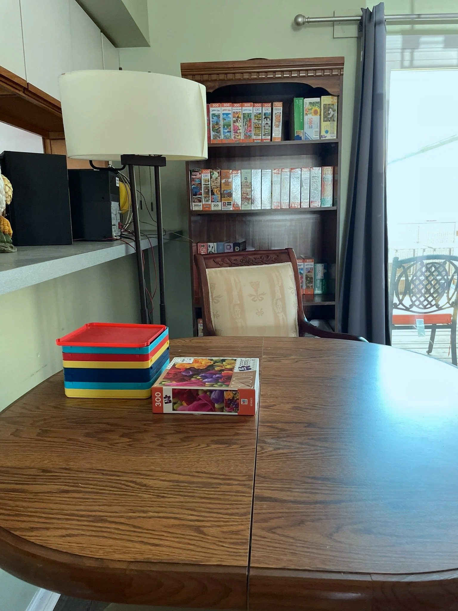 A wooden dining table with a colorful stacking tray and a seed packet featuring pink and purple flowers. In the background, there's a bookshelf filled with puzzles or books, a chair with a patterned cushion, a standing lamp, and a sliding glass door 