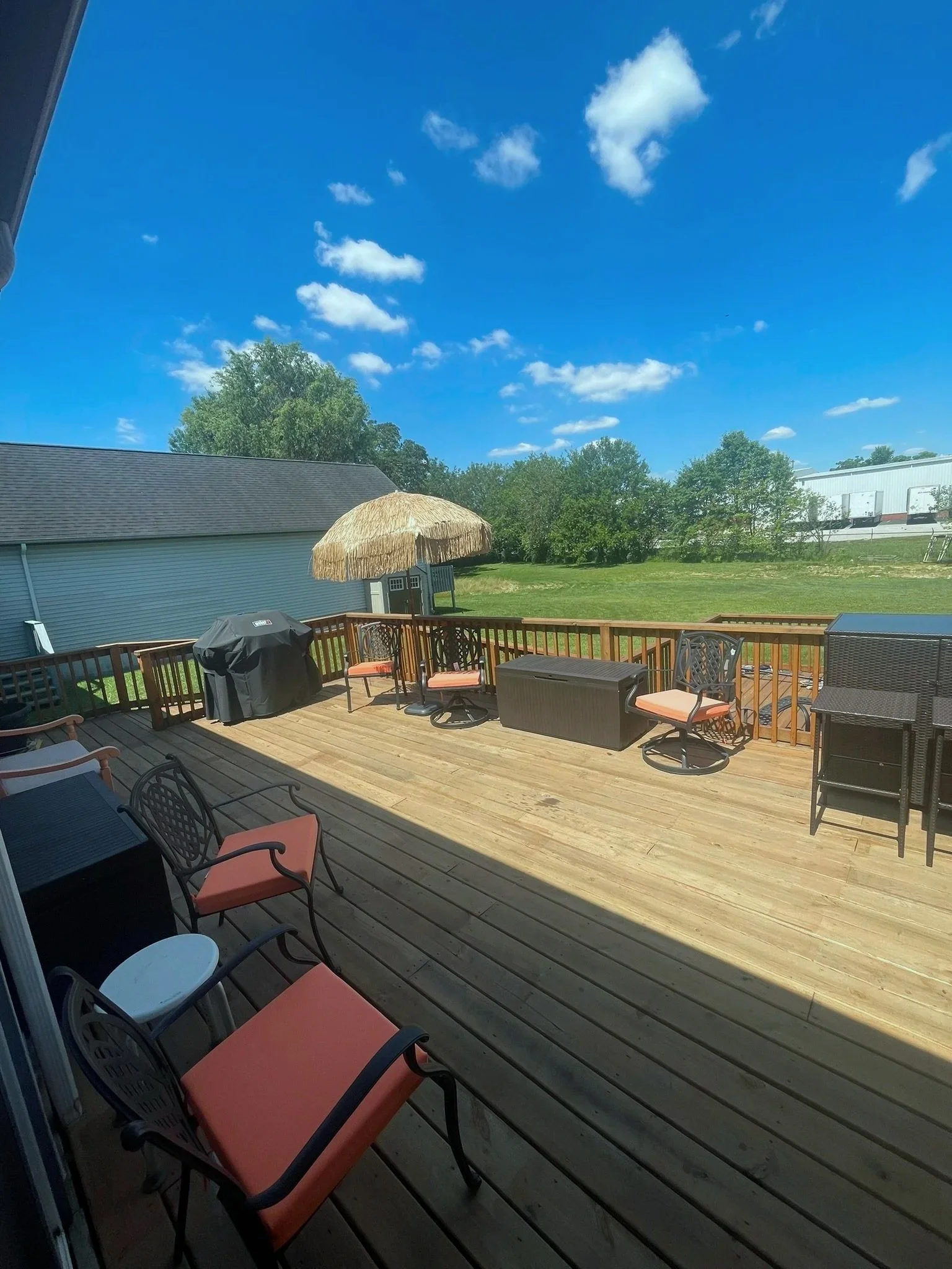 A wooden deck with outdoor furniture, a grill, a straw umbrella, and a grassy backyard with trees and a clear blue sky with clouds.