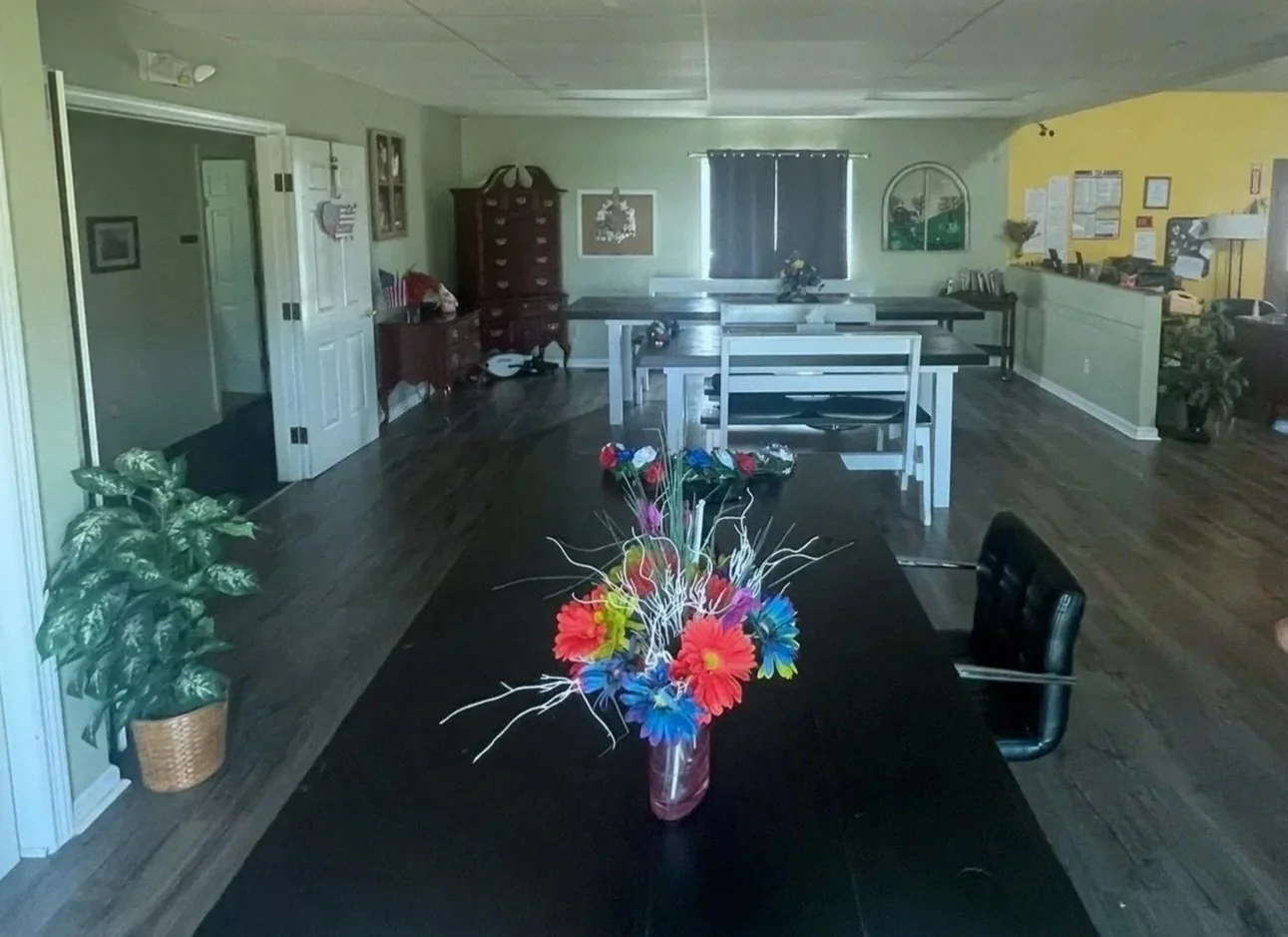 Interior of a spacious room with a black rectangular table in the foreground, decorated with a vase of colorful artificial flowers. The room features a wooden floor, light green walls, and a ceiling with white tiles. In the background, there are vari