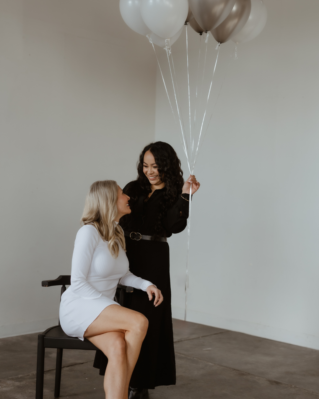 Two women celebrating with balloons, one seated in a wheelchair, smiling at each other in a minimalist room.