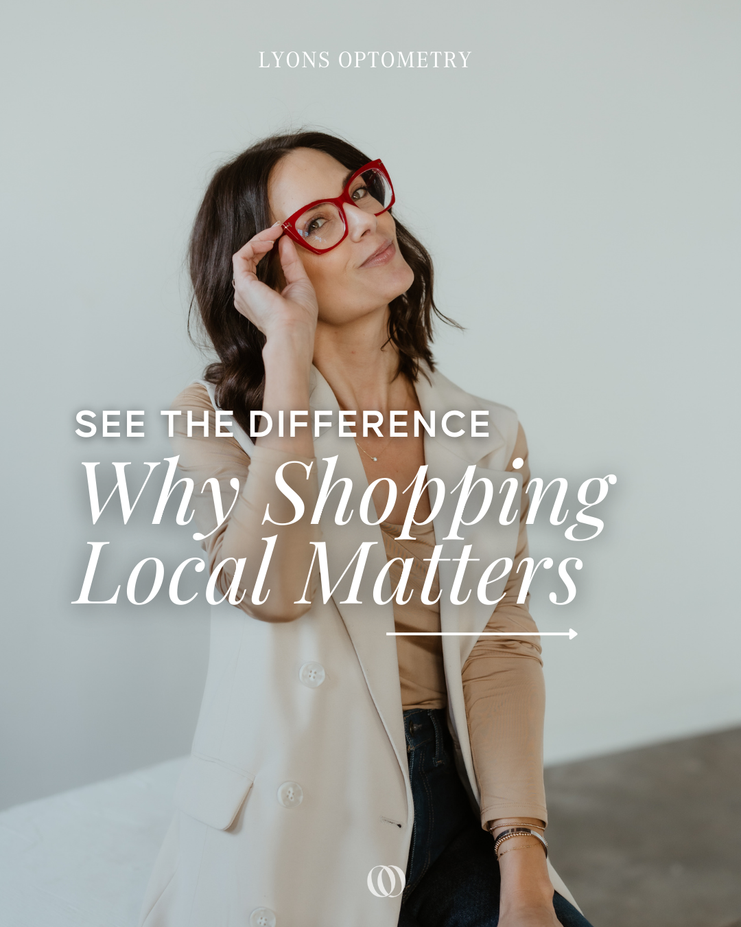A woman with long dark hair wearing red glasses and a beige blazer, looking thoughtfully at the camera with one hand adjusting her glasses.