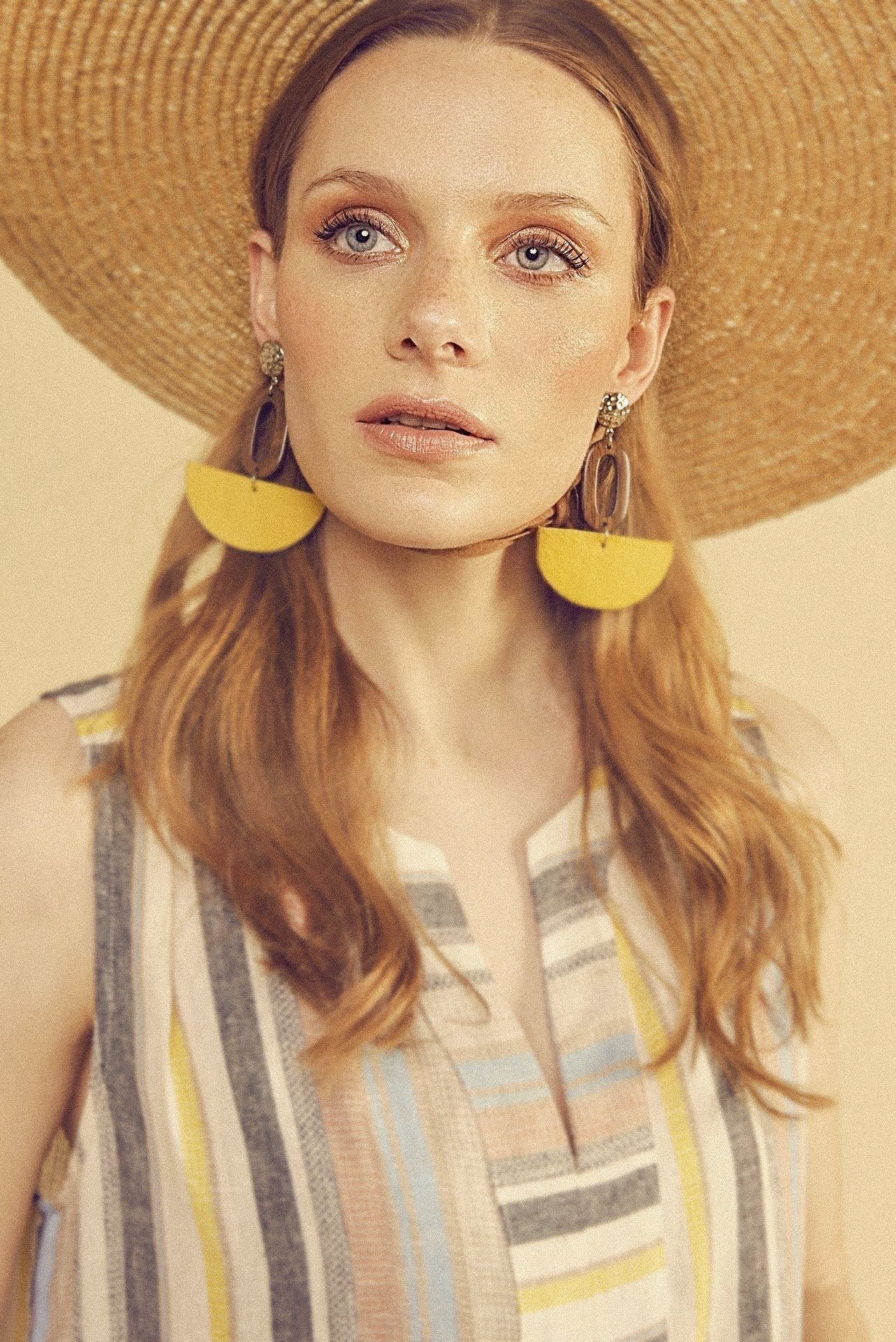A woman with long red hair wearing a large straw hat, gold and purple earrings, and a sleeveless dress with multicolored vertical stripes.