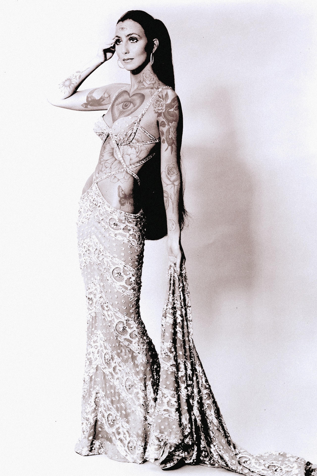 A woman with multiple tattoos, wearing a sheer, embroidered gown, standing against a plain white background.