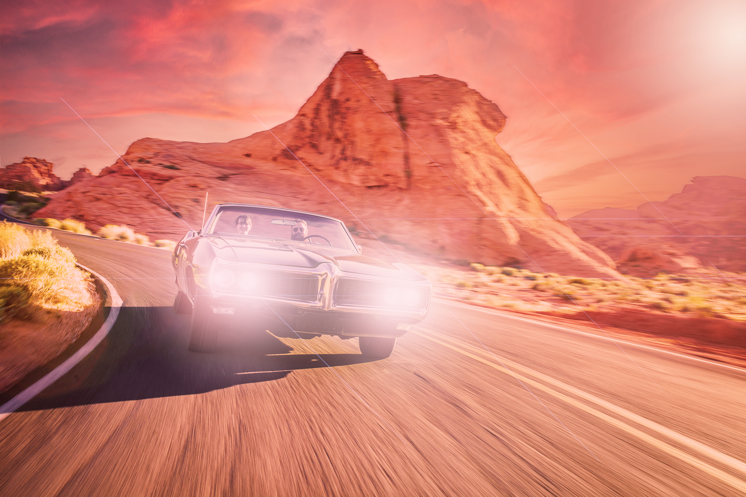 A vintage car with headlights on, driving on a winding road through a desert landscape with red rock formations and mountains under a pink and orange sunset sky.