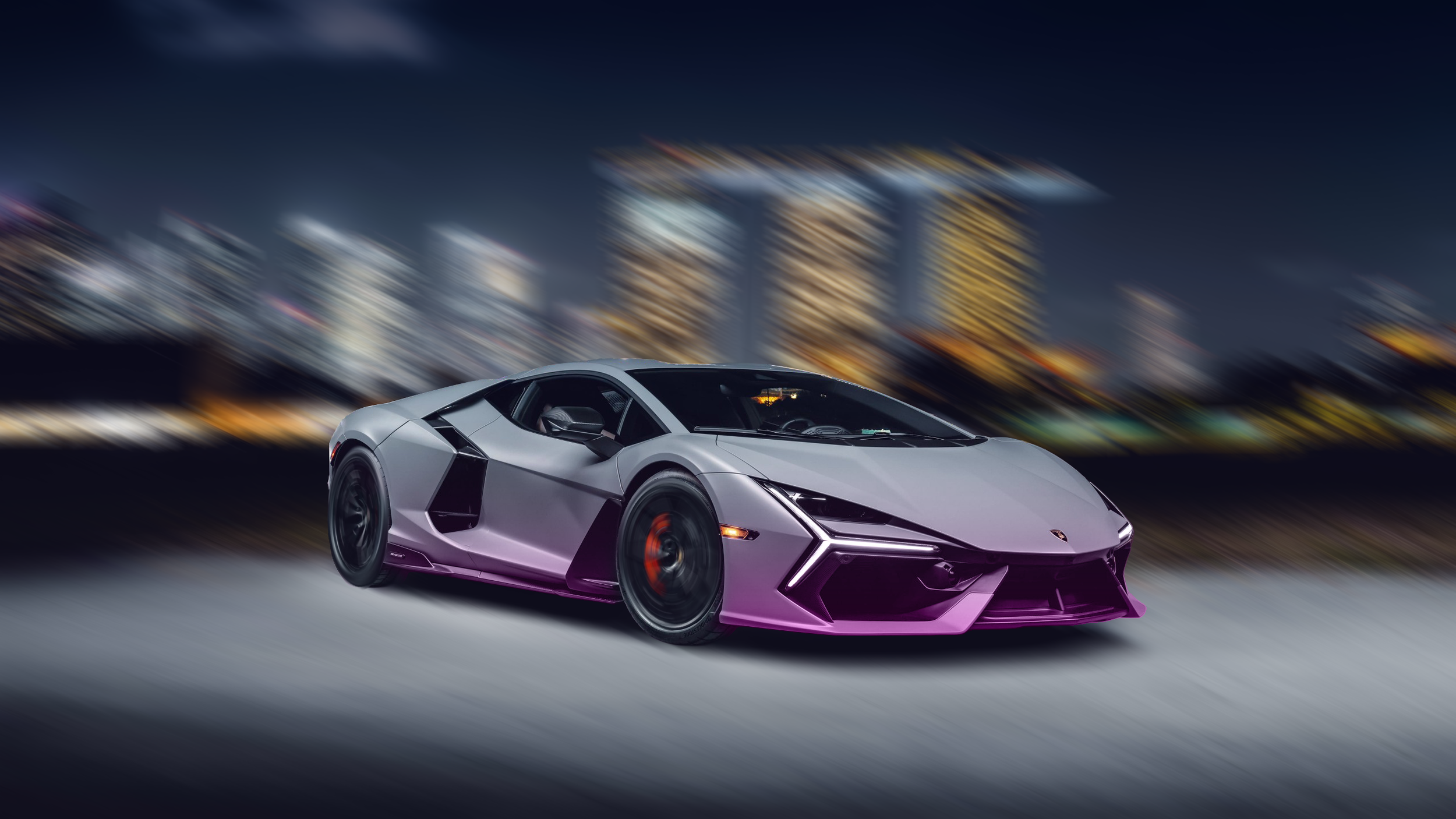 A silver Lamborghini sports car driving at night with blurred city lights in the background.