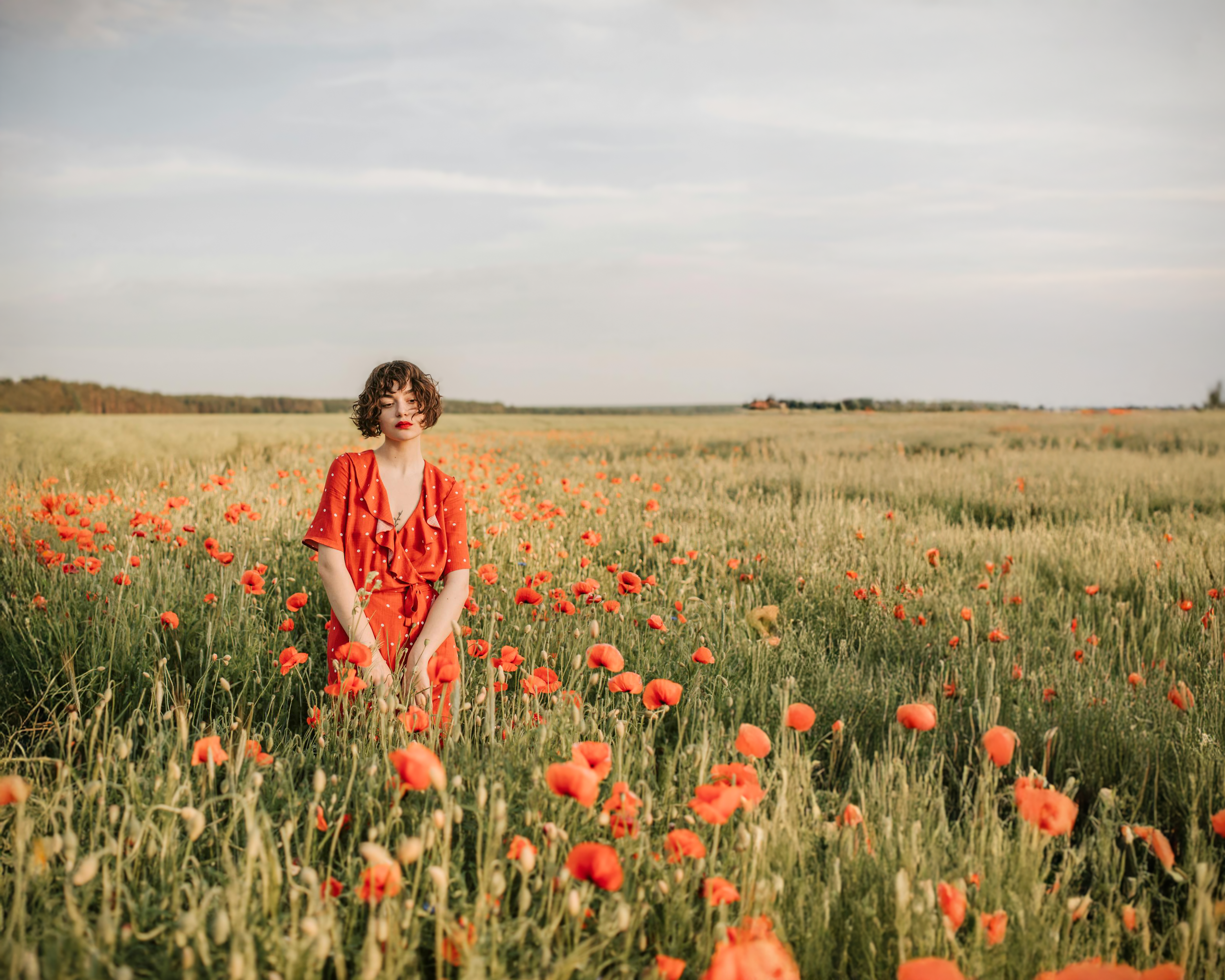 A woman in a red dress standing in a field of orange poppies under a cloudy sky.
