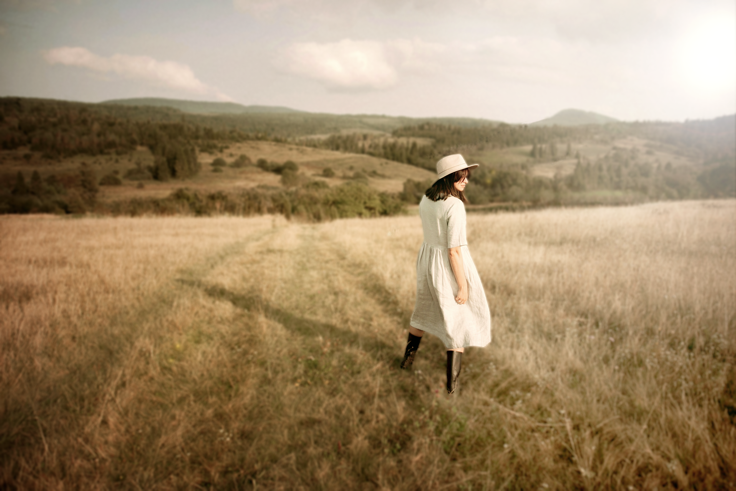 A woman wearing a white dress, black boots, and a hat walking through a grassy field with rolling hills and distant trees in the background.