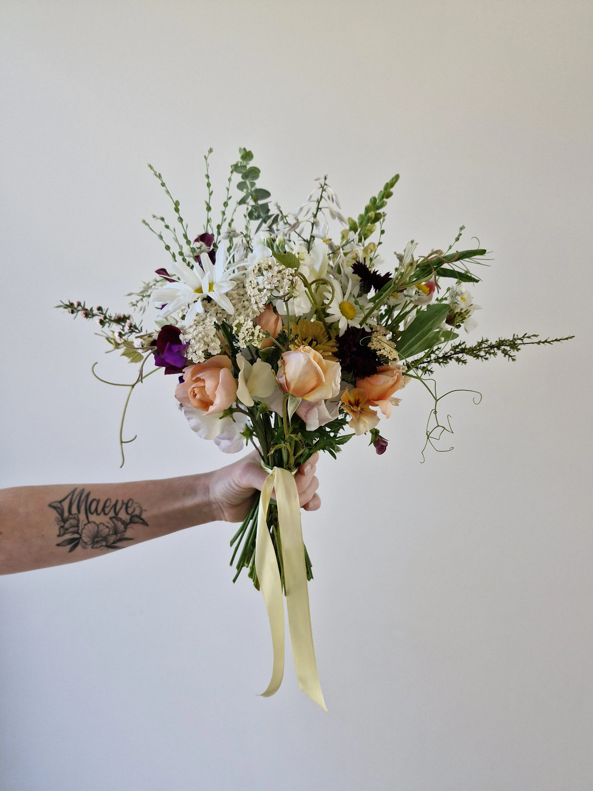 Bride holding a colorful bouquet of flowers with a yellow ribbon against a plain wall.
