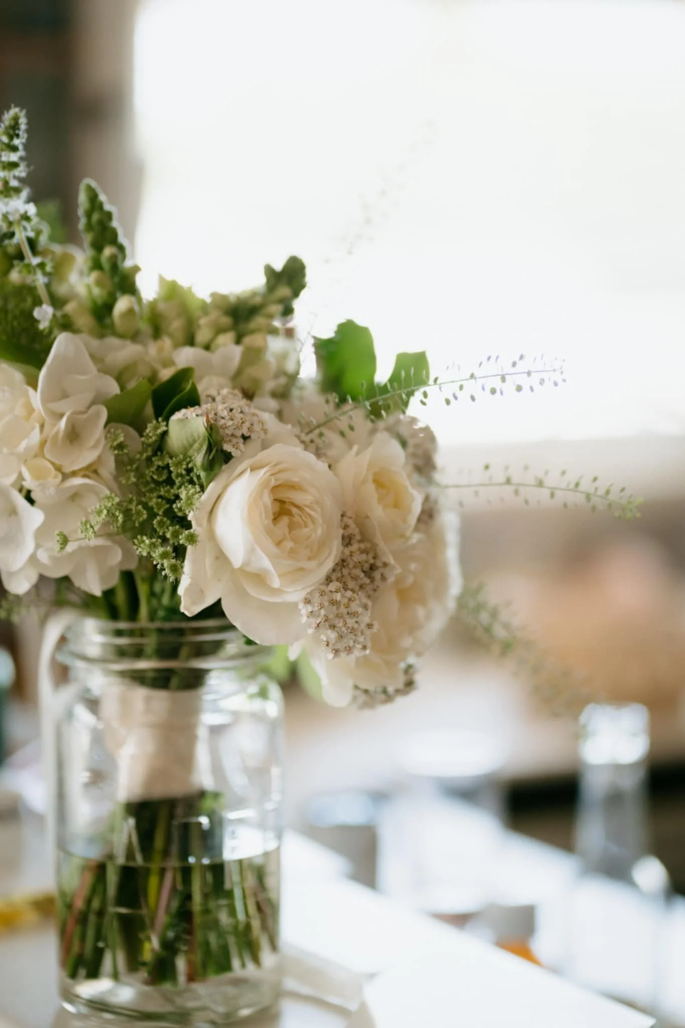 White floral wedding bouquet in a glass jar on a table, featuring roses, and hydrandeas, the bride has left her bouquet on the bridal table.