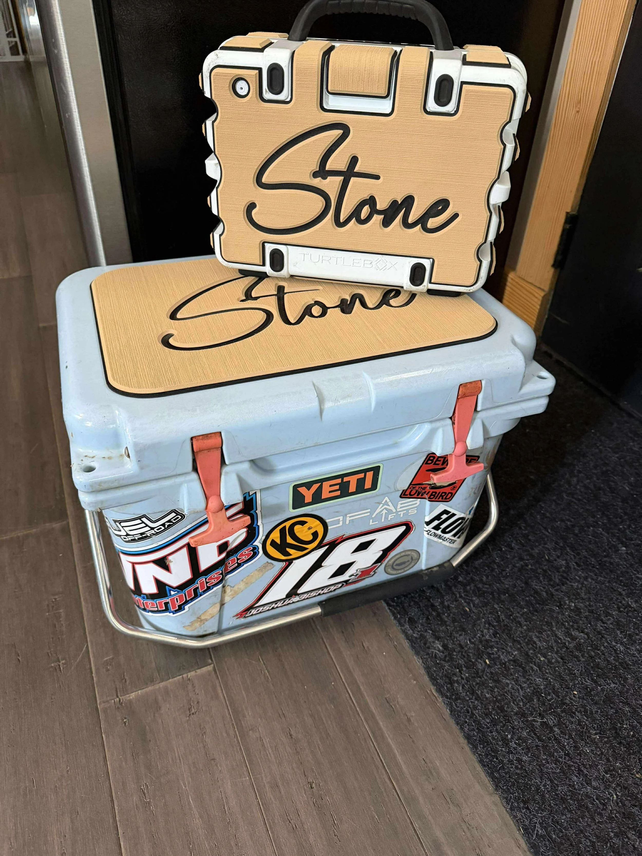 A white cooler decorated with various stickers and two orange clips. On top of it are two wooden signs with black lettering spelling 'Stone.'