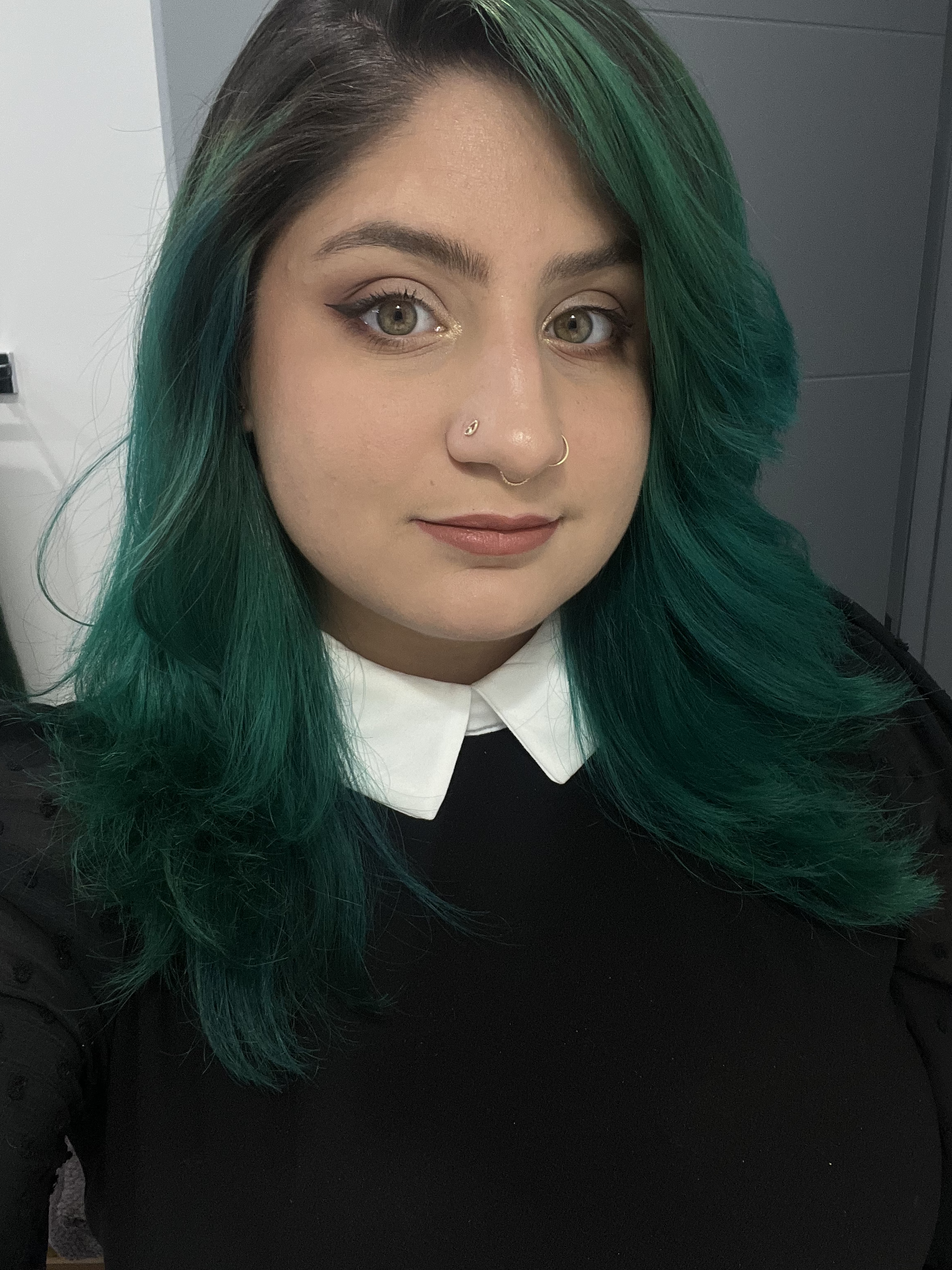 Close-up of a woman with green hair, wearing a black top with a white collar, and makeup featuring eyeliner and neutral lipstick.