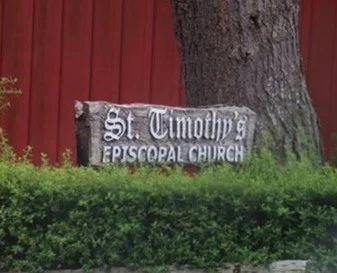 Image of a wood sign behind a green hedge reads St Timothy's Episcopal Church in white, red building and tree behind the sign.