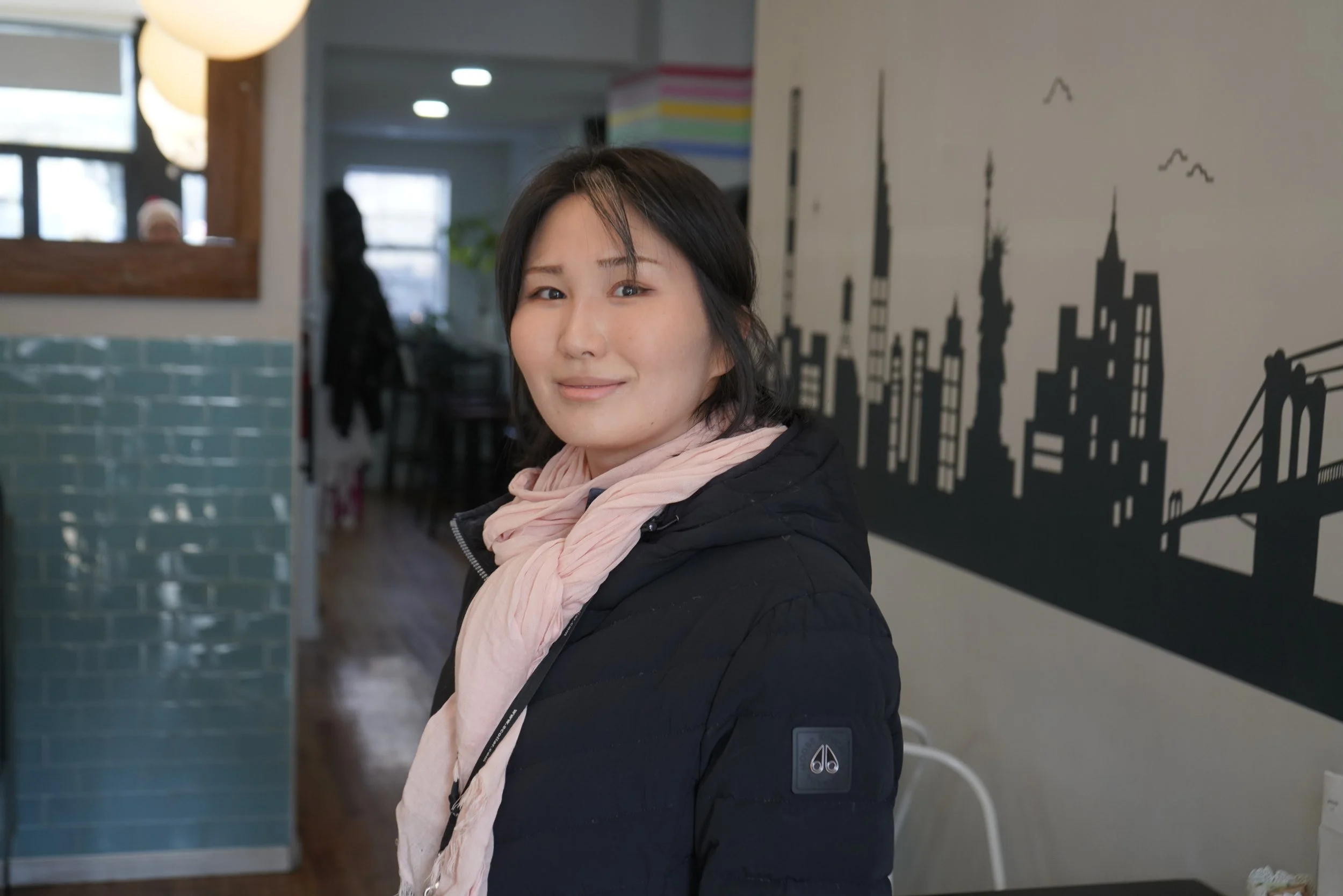 Psychic medium Cherry Sees with shoulder-length black hair standing indoors, smiling slightly, wearing a black jacket and a light pink scarf. Behind her is a cityscape silhouette wall art and a window with natural light.