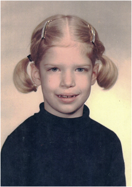 A young girl with blonde hair styled in pigtails, wearing a dark top, and looking at the camera.