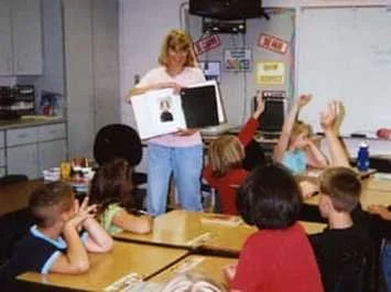 Teacher reading a book to a classroom of students raising their hands.