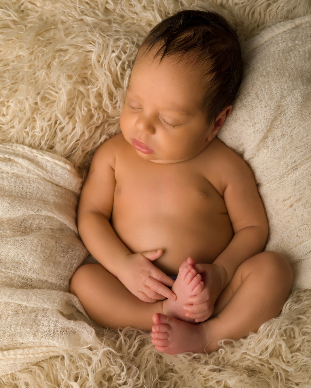 A sleeping baby with dark hair, laying on a fluffy cream-colored blanket with a pillow, resting peacefully.