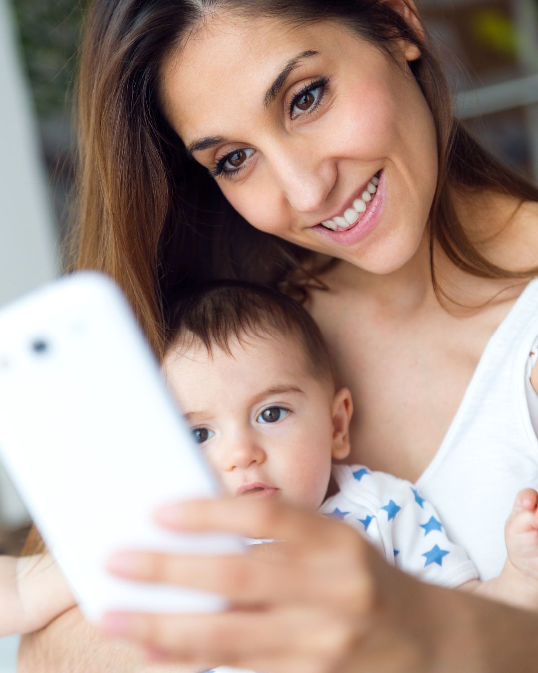 A young woman with long brown hair smiling and taking a selfie with her toddler, who is looking at the phone with a curious expression, both close together.