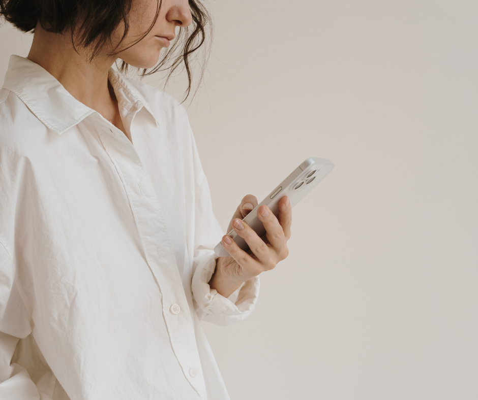 A woman with shoulder-length dark hair wearing a white button-up shirt looking at a smartphone in her hand against a plain, off-white background.