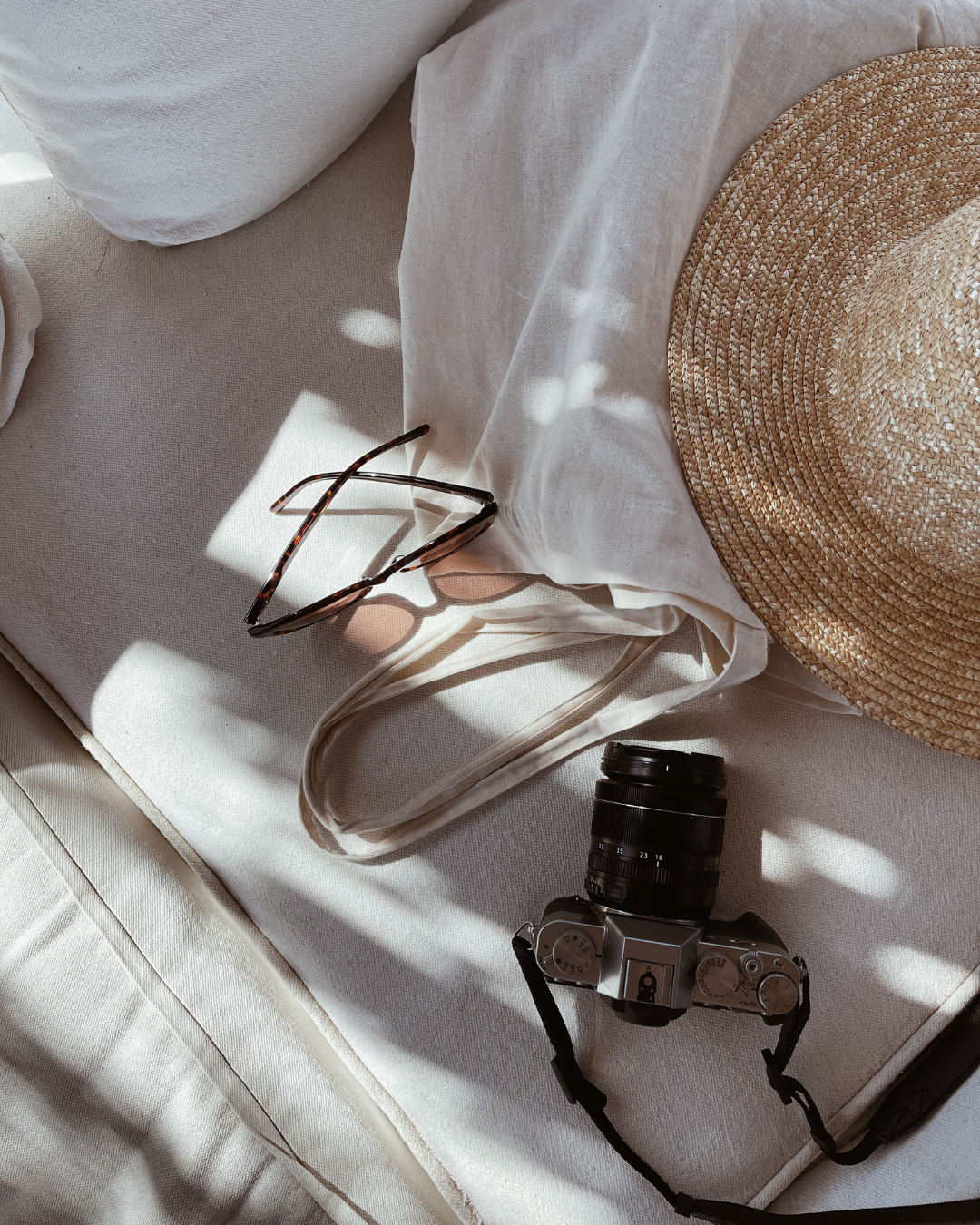 A photograph of a light-colored fabric surface with a straw hat, a pair of tortoiseshell sunglasses, a camera, and a white tote bag with handles. Sunlight and shadows are visible on the objects.