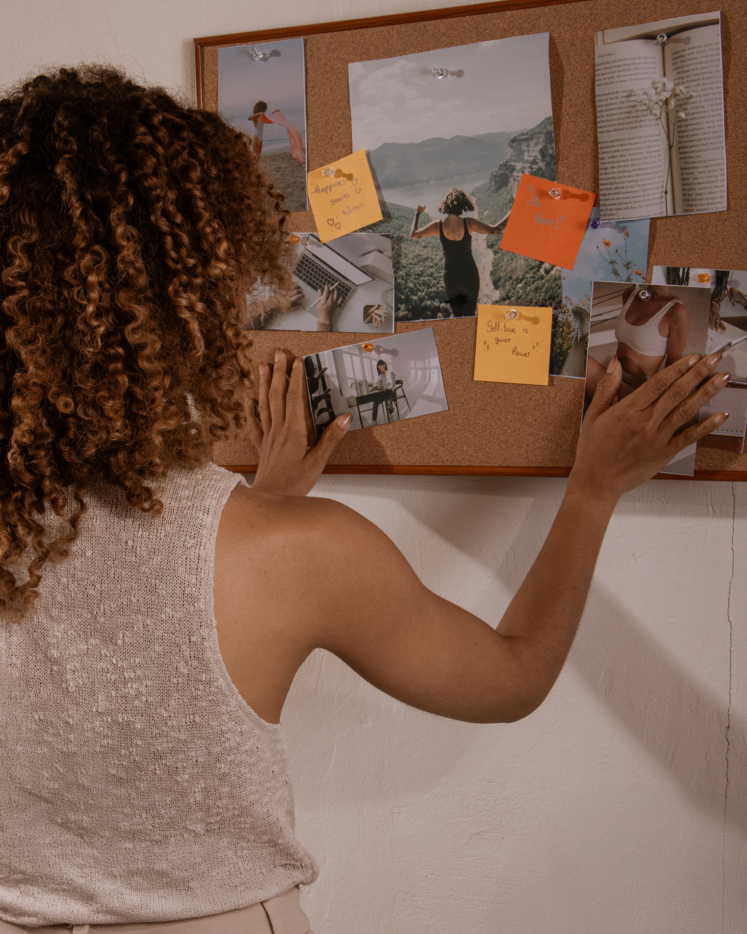 A woman with curly hair pins photos and notes on a corkboard, with some images of landscapes, a person outdoors, and a person working at a desk.