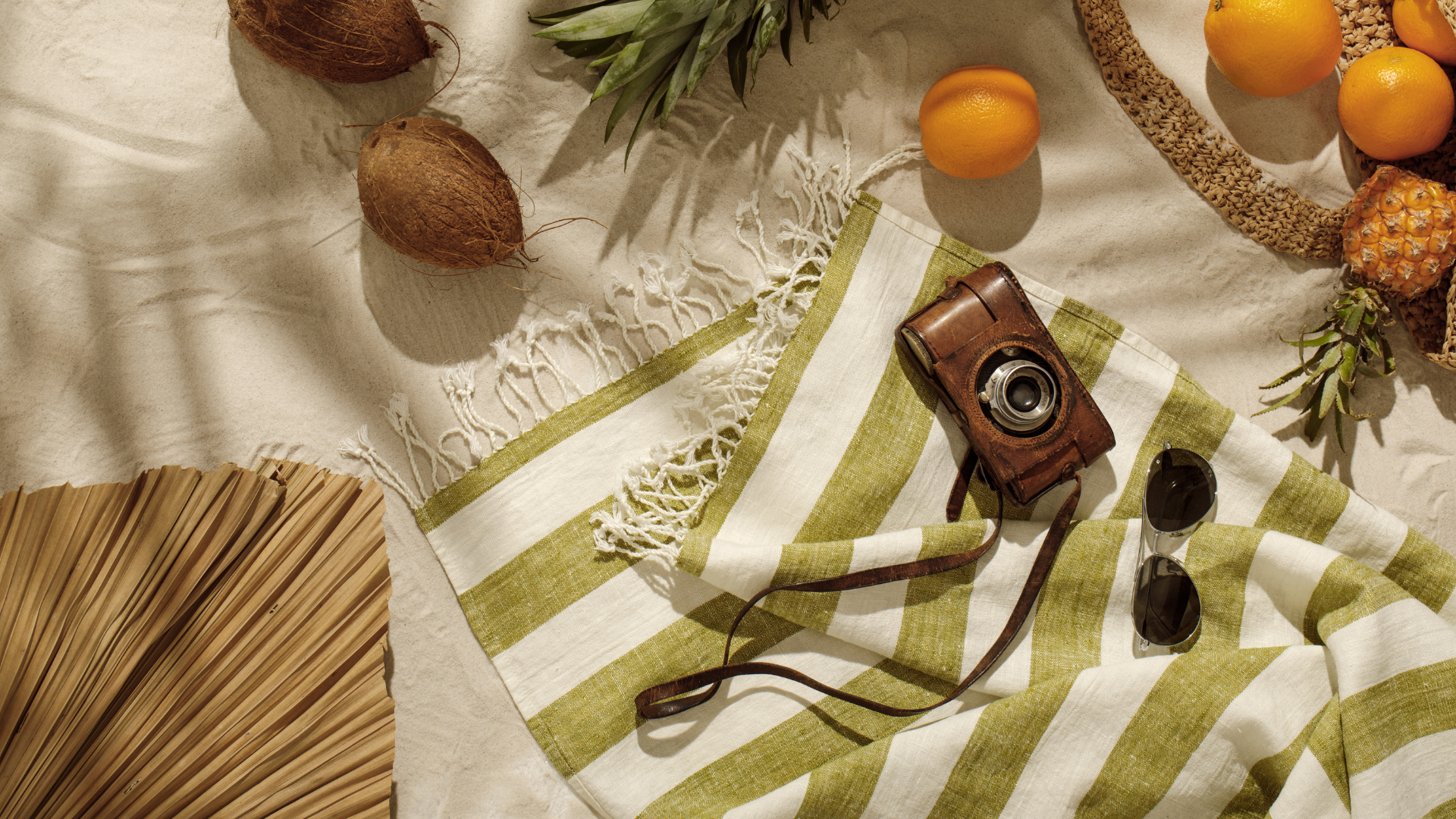 A collection of tropical fruits, including coconuts, pineapples, oranges, and a pineapple, on a beige fabric background, alongside a vintage camera, sunglasses, a woven bag, and a striped green and white cloth.