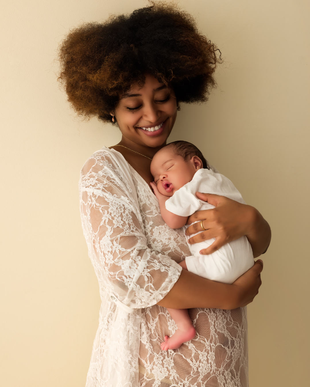 A smiling woman with curly hair holding a sleeping baby in white clothing