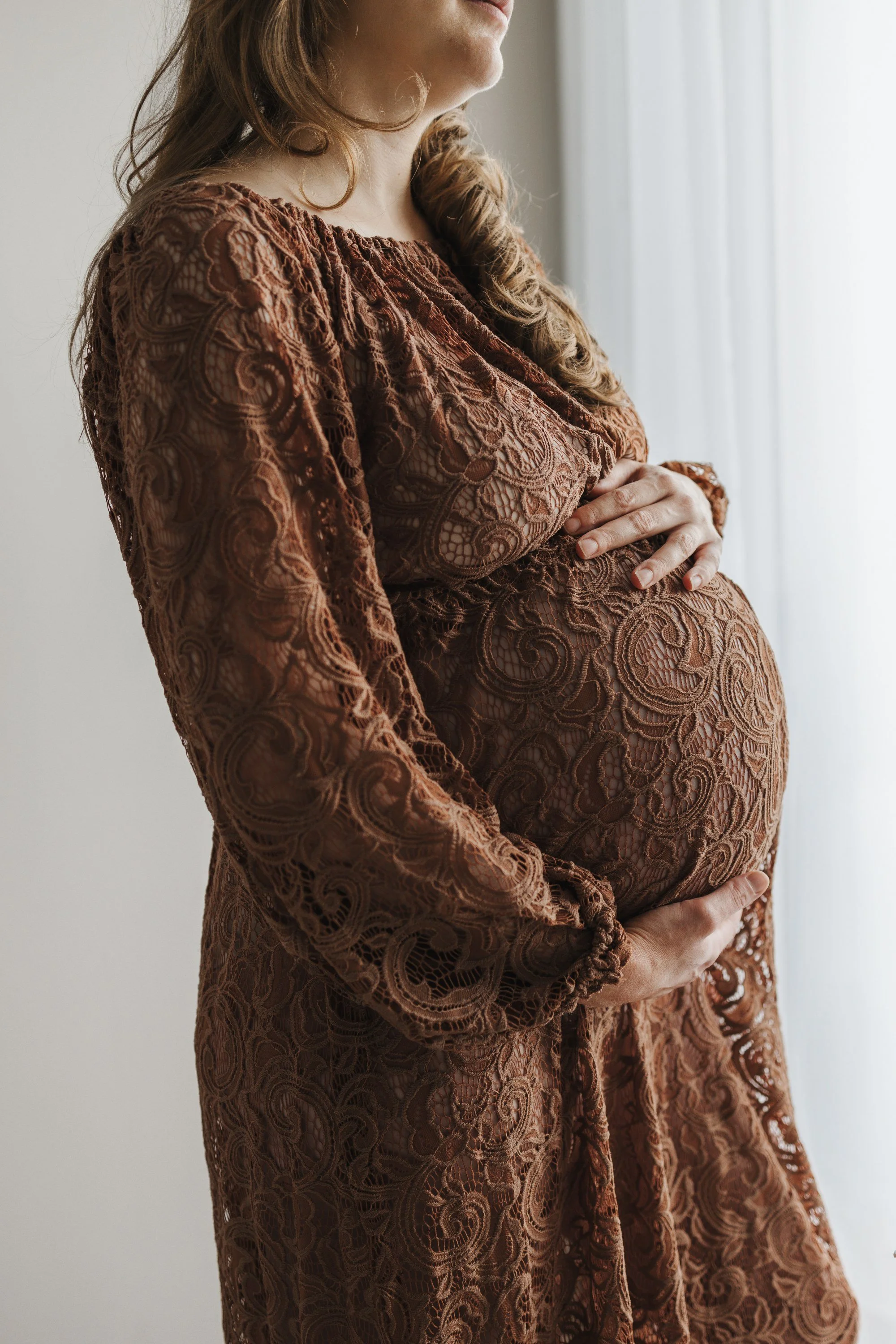 A pregnant woman in a brown lace dress is cradling her belly with her hands, standing near a window with white curtains.