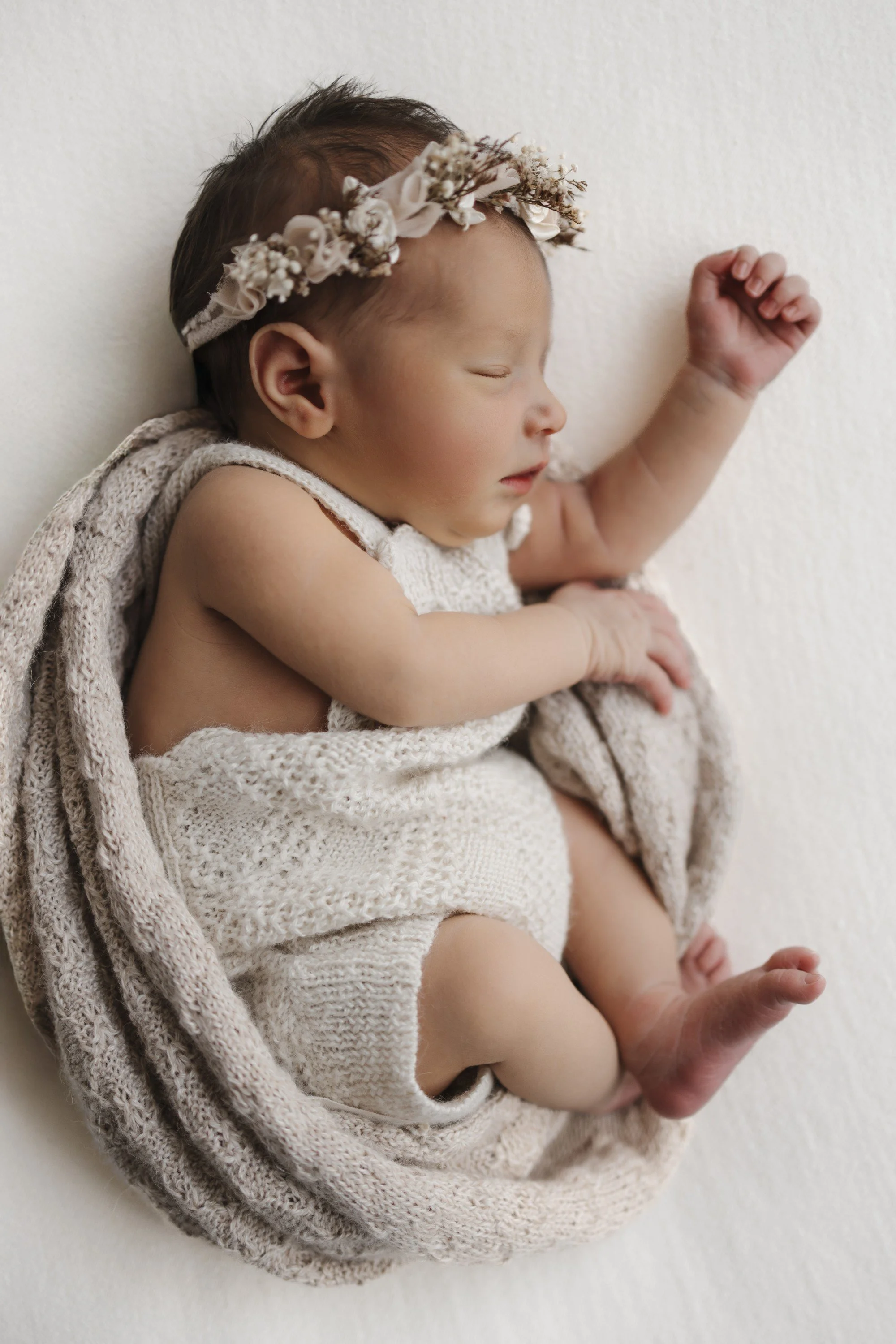 A sleeping baby girl with a floral headband, wrapped in a cream knitted blanket, lying on a white surface.