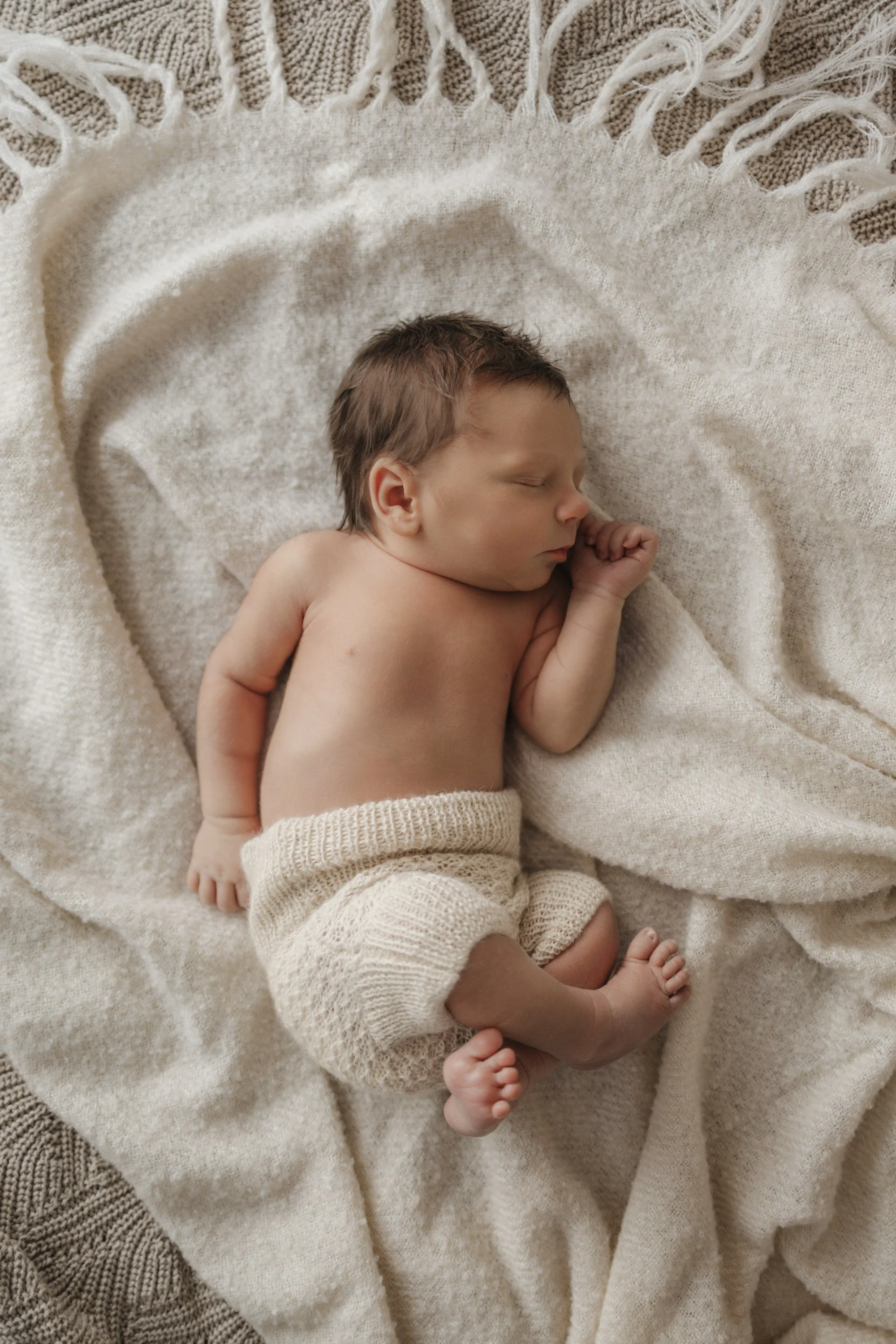 A sleeping newborn baby lies on a soft, cream-colored blanket, wearing knitted cream shorts, with eyes closed and arms gently curled.