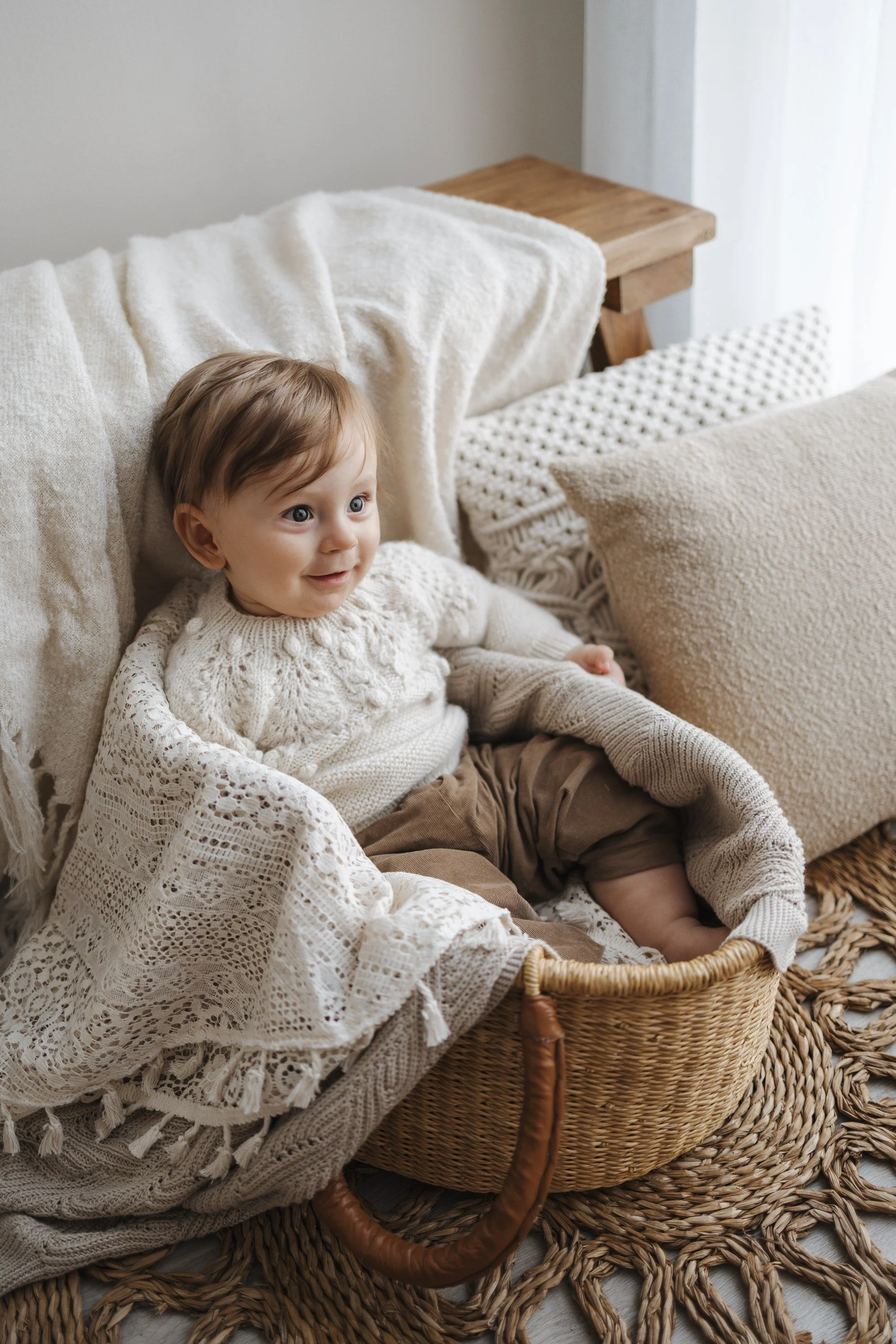 A young child with brown hair and blue eyes sitting in a woven basket, wearing a cream knit sweater and brown pants, surrounded by soft blankets and pillows in a cozy living room.