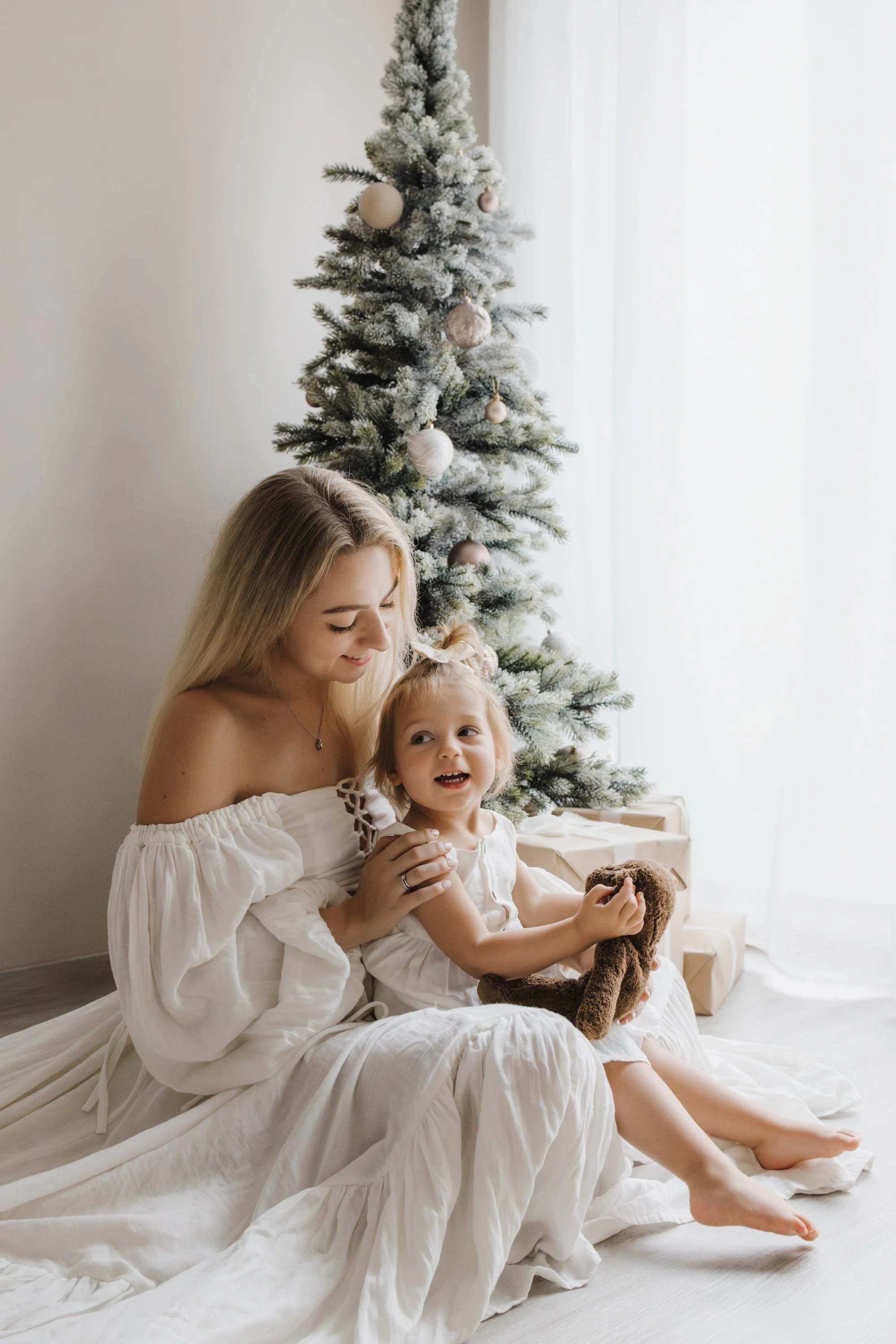 A woman and a young girl sitting on the floor near a decorated Christmas tree with wrapped gifts, both smiling and holding a stuffed animal, dressed in white, with natural light coming from a window.