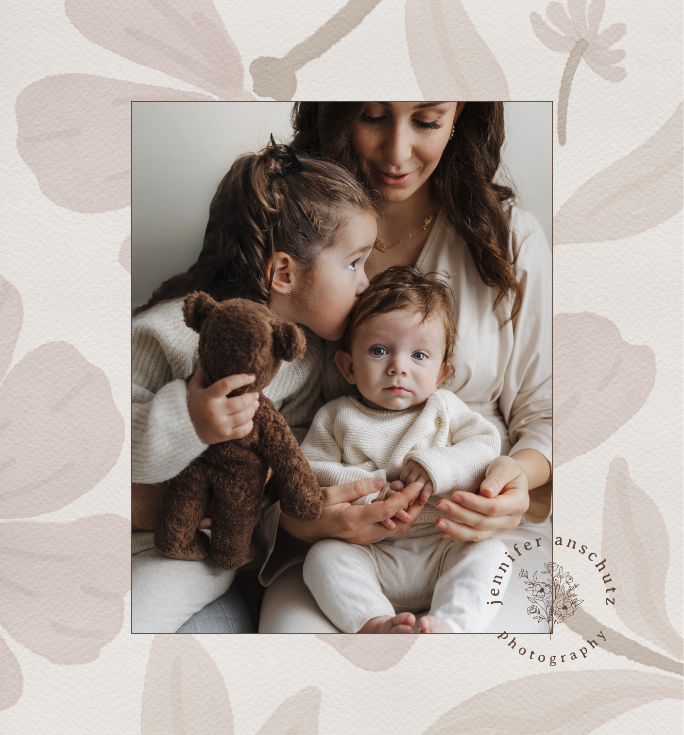 A woman with two young children, one girl and one boy, sitting closely together indoors. The girl is kissing the boy's head while holding a brown teddy bear, and the woman is holding the boy's hand. The children are dressed in light-colored clothing, and the background features a decorative floral pattern with a signature and logo of the photographer, Jennifer Franschutz.