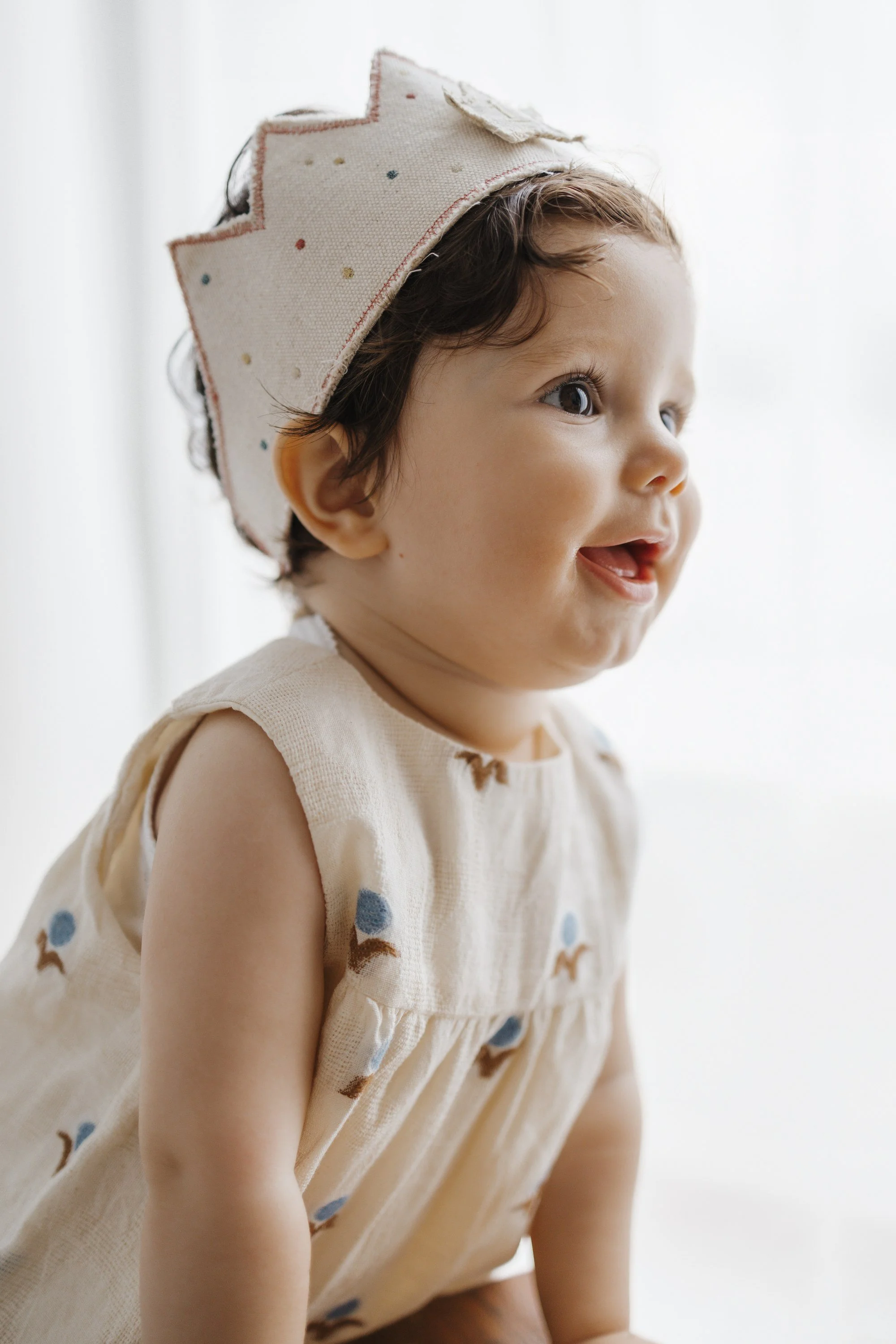 A smiling toddler with dark hair and brown eyes, sitting on the floor wrapped in a sheer white curtain.