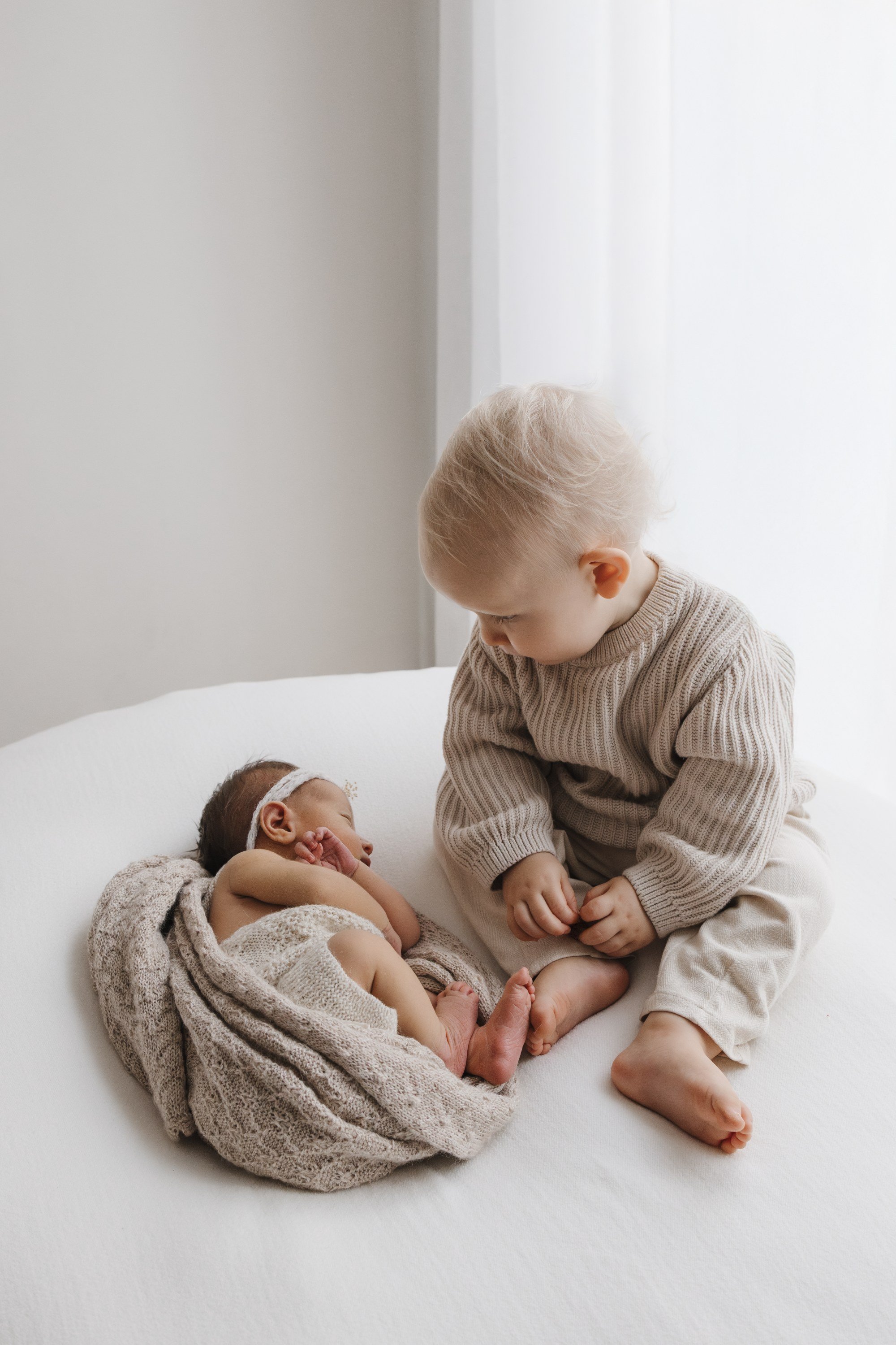 A young boy sitting on a white bed, looking at a sleeping newborn baby who is wrapped in a beige knitted blanket.