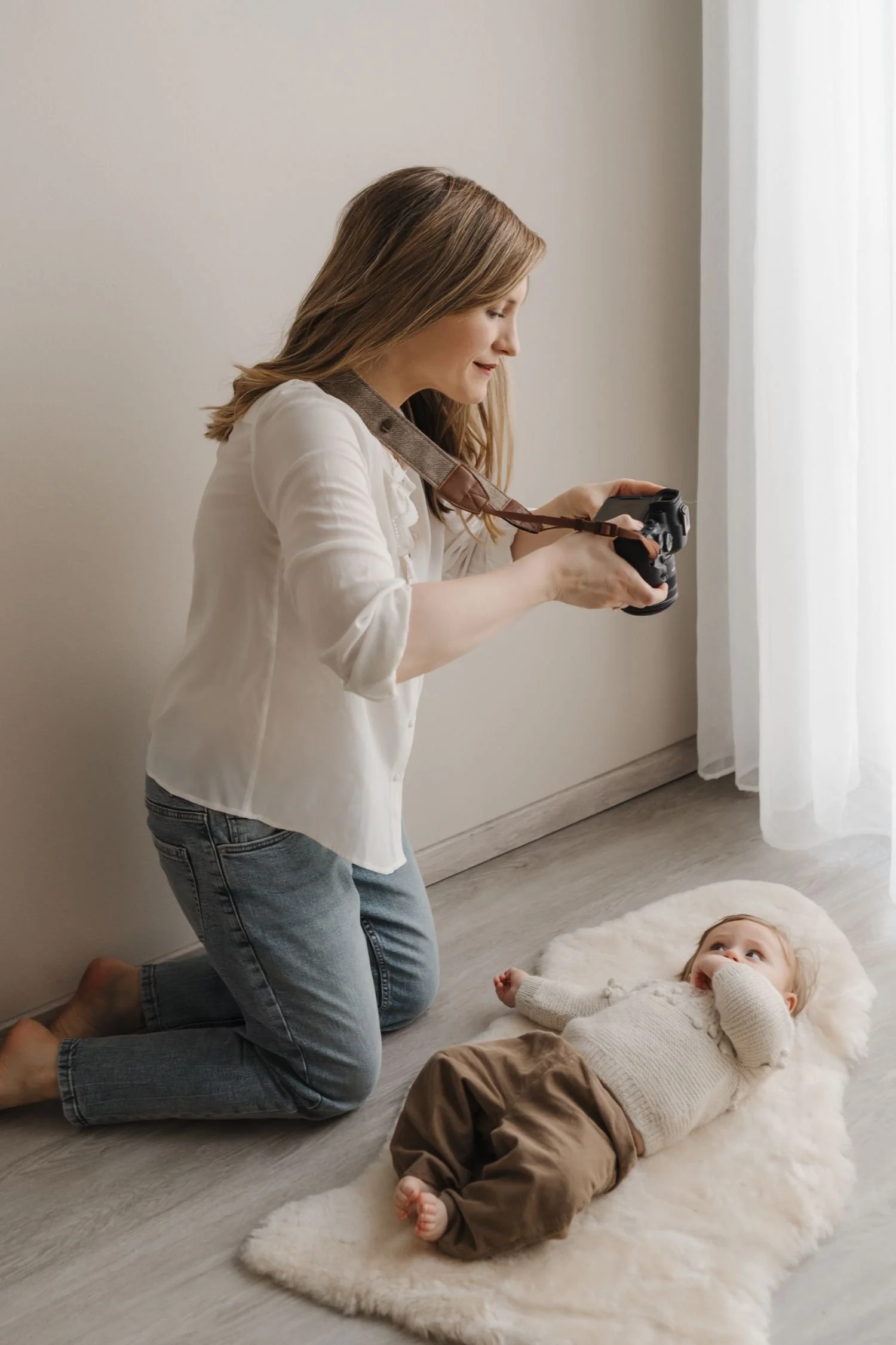 A woman kneeling on the floor taking a photo of a young child lying on a plush rug, with sunlight coming through a window.