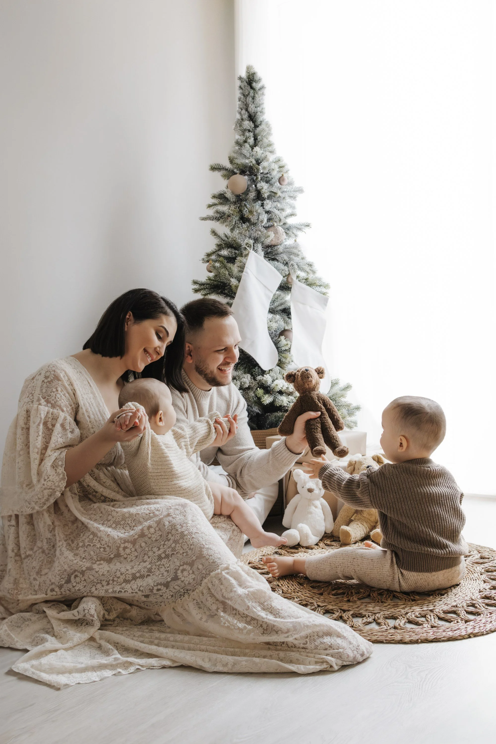 A woman holding a baby, with a man leaning in from the top, in a cozy, neutral-toned setting. The baby looks directly at the camera, holding a small wooden block.