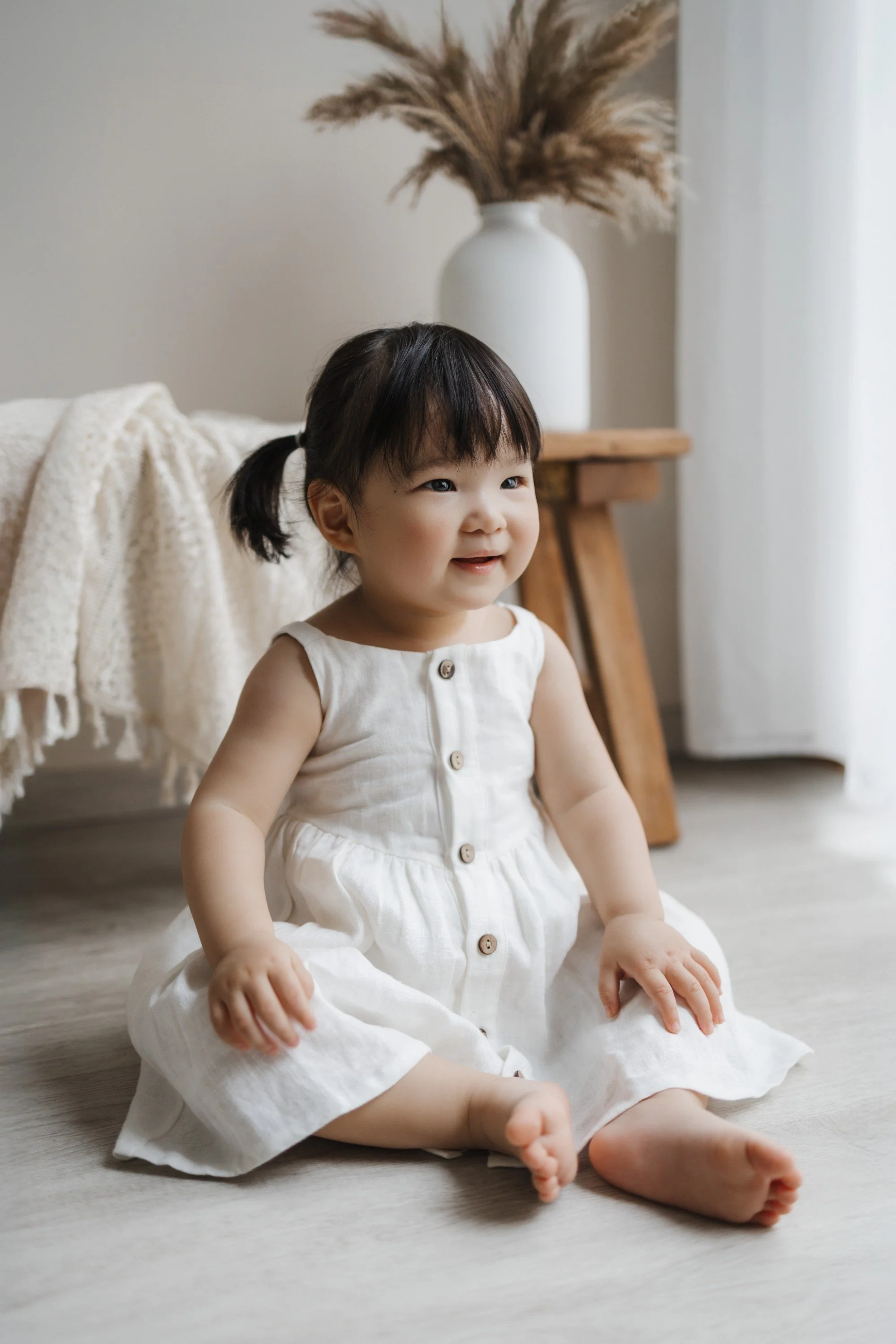 A smiling toddler girl sitting on the floor in a white dress, with a cozy blanket and a vase with pampas grass in the background.