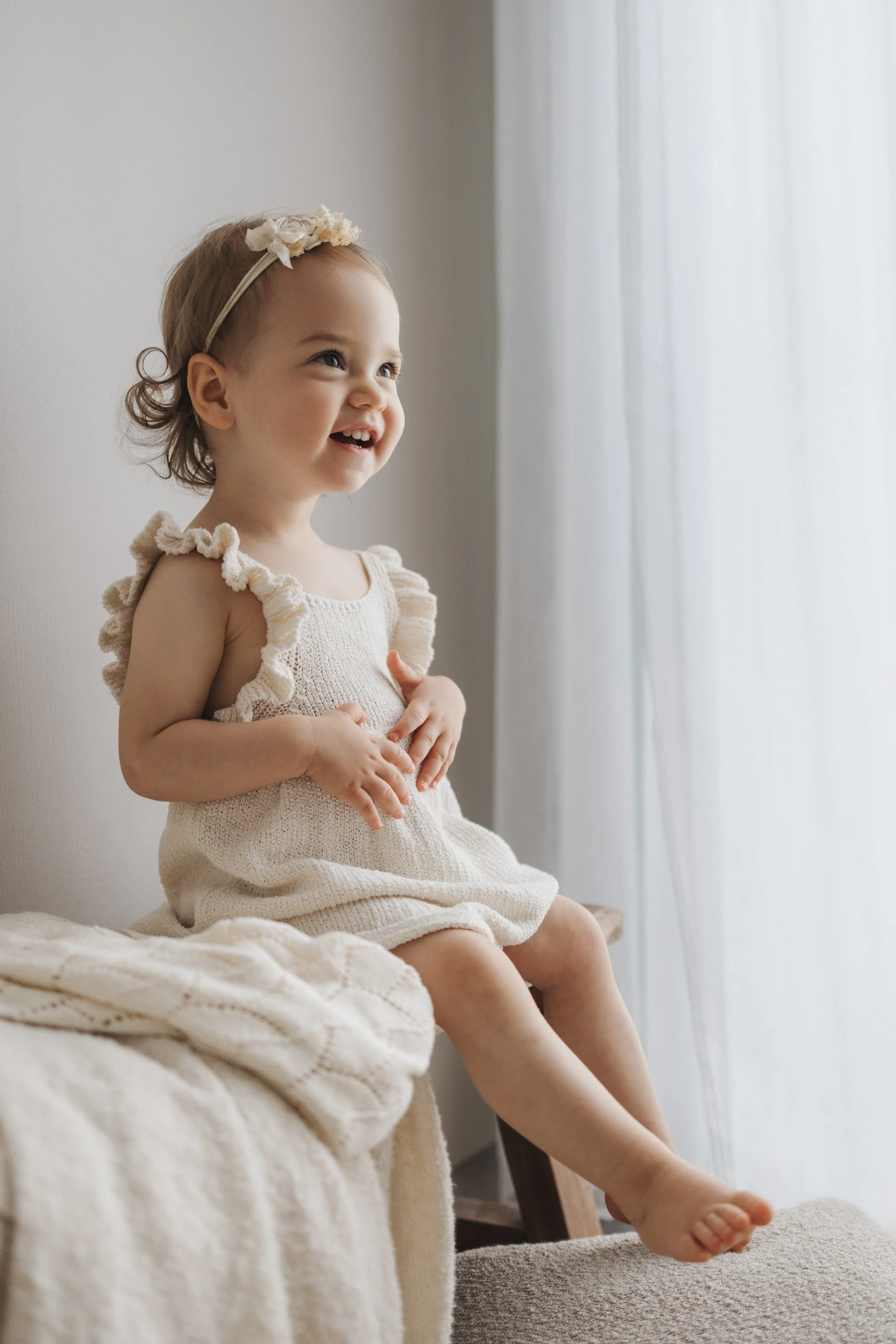 A young girl with a flower headband and a white dress sitting on a blanket-covered surface near a window, smiling and looking to the side.